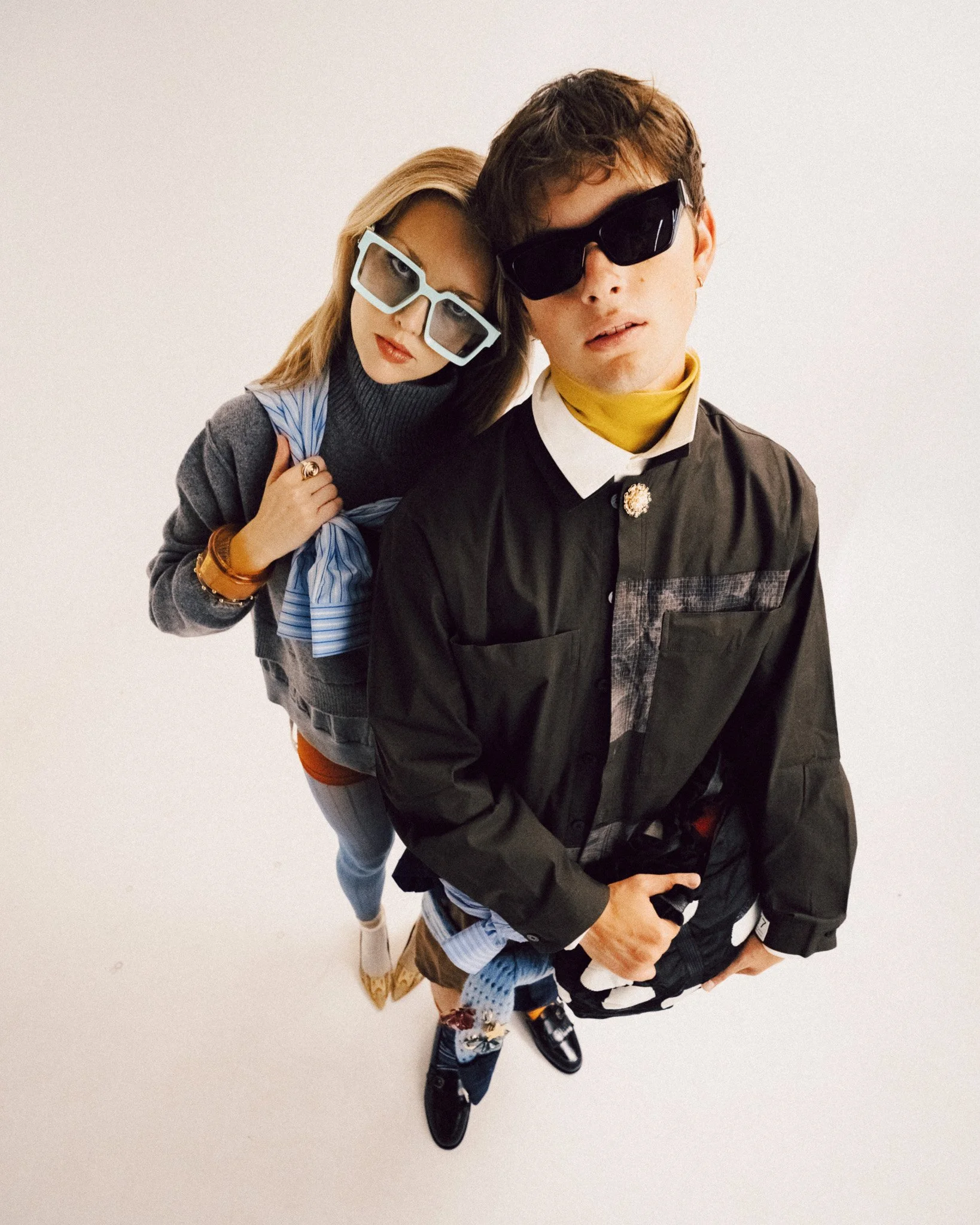 A young woman and man posing together against a plain background, wearing modern layered streetwear and oversized sunglasses