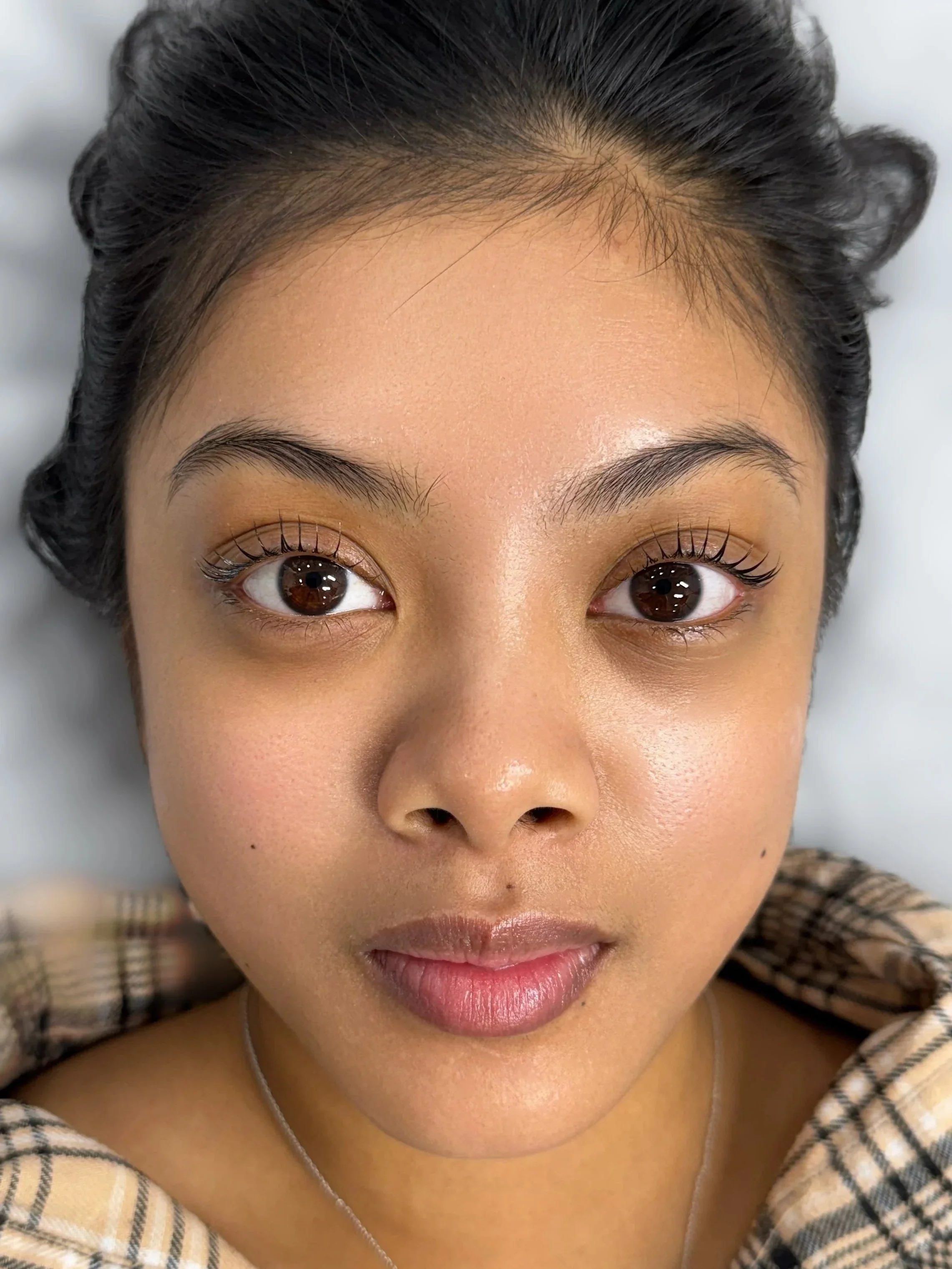 A woman with dark hair, styled with volume, looking directly at the camera, wearing a plaid shirt and a necklace, with makeup including eyeliner, mascara, and natural pink lipstick.