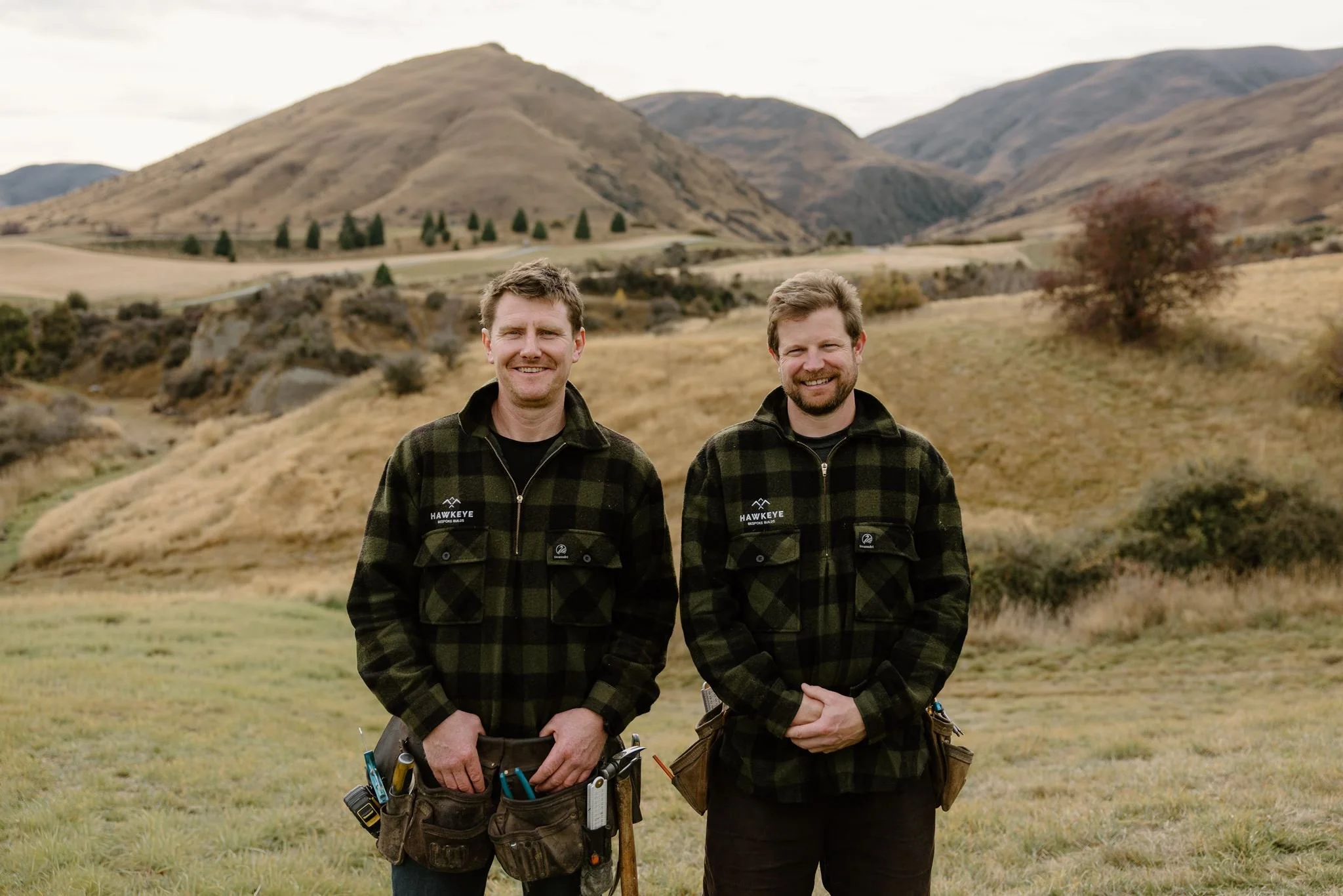 Two men standing side by side outdoors in a hilly landscape, wearing matching black and green plaid jackets with the 'Hawkeye' logo, smiling at the camera.