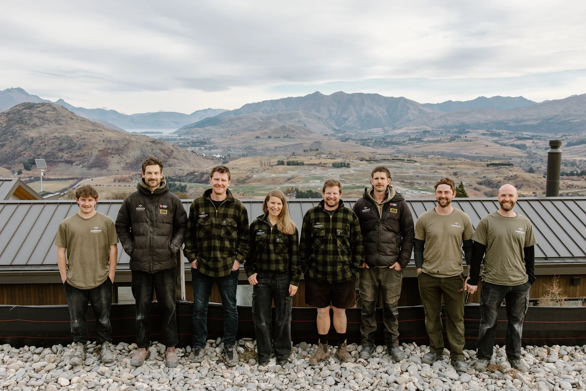 A group of nine people standing on rocks in front of a mountain landscape with cloudy skies.