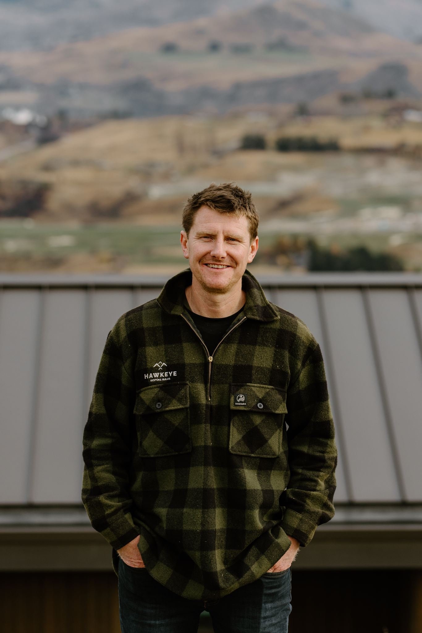 A man with short brown hair and a beard, smiling, wearing a dark green and black plaid jacket with patches, standing outdoors in front of blurred landscape hills and trees.
