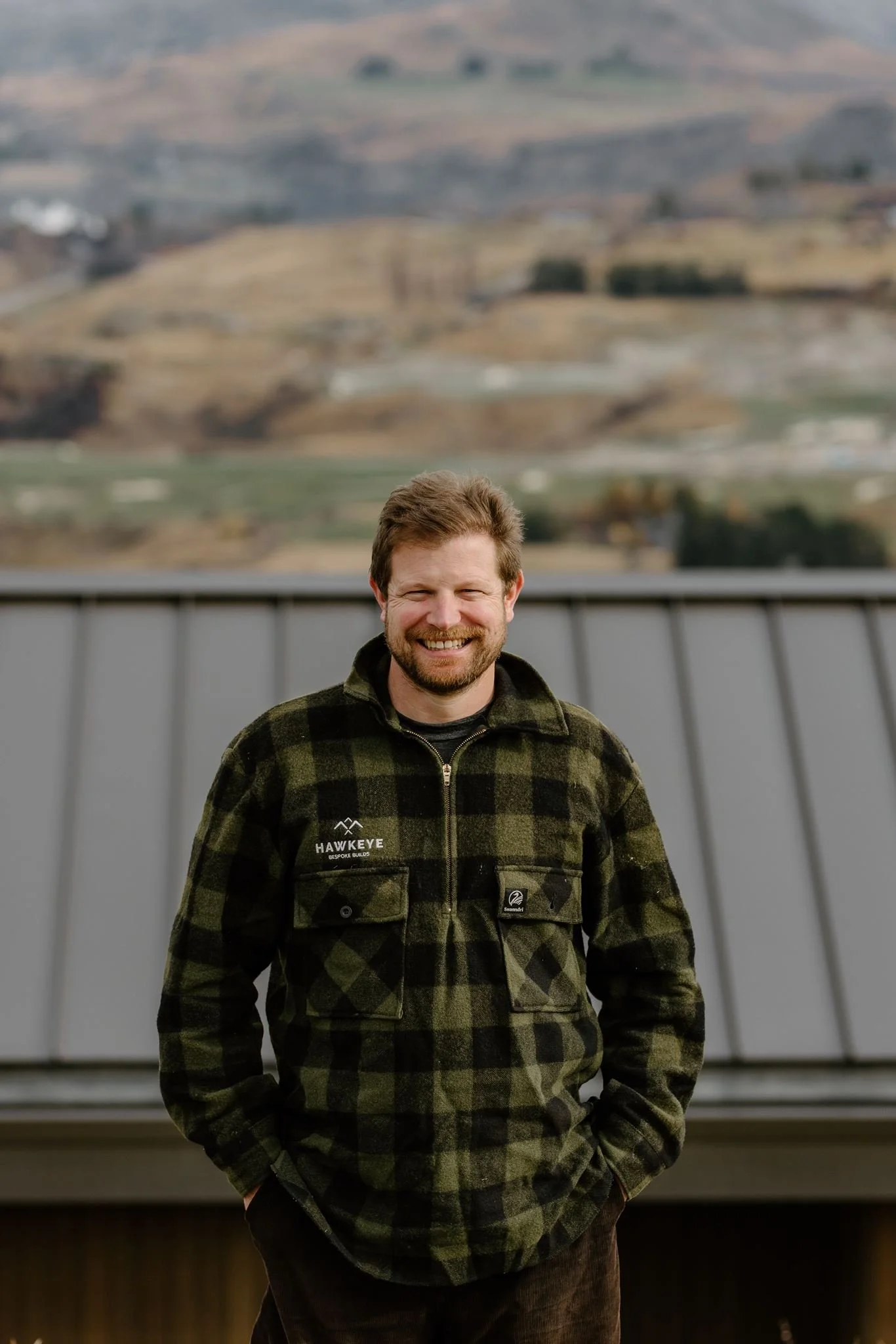 A smiling man with a beard wearing a green and black checkered jacket standing outdoors with a blurred landscape of rolling hills and fields in the background.