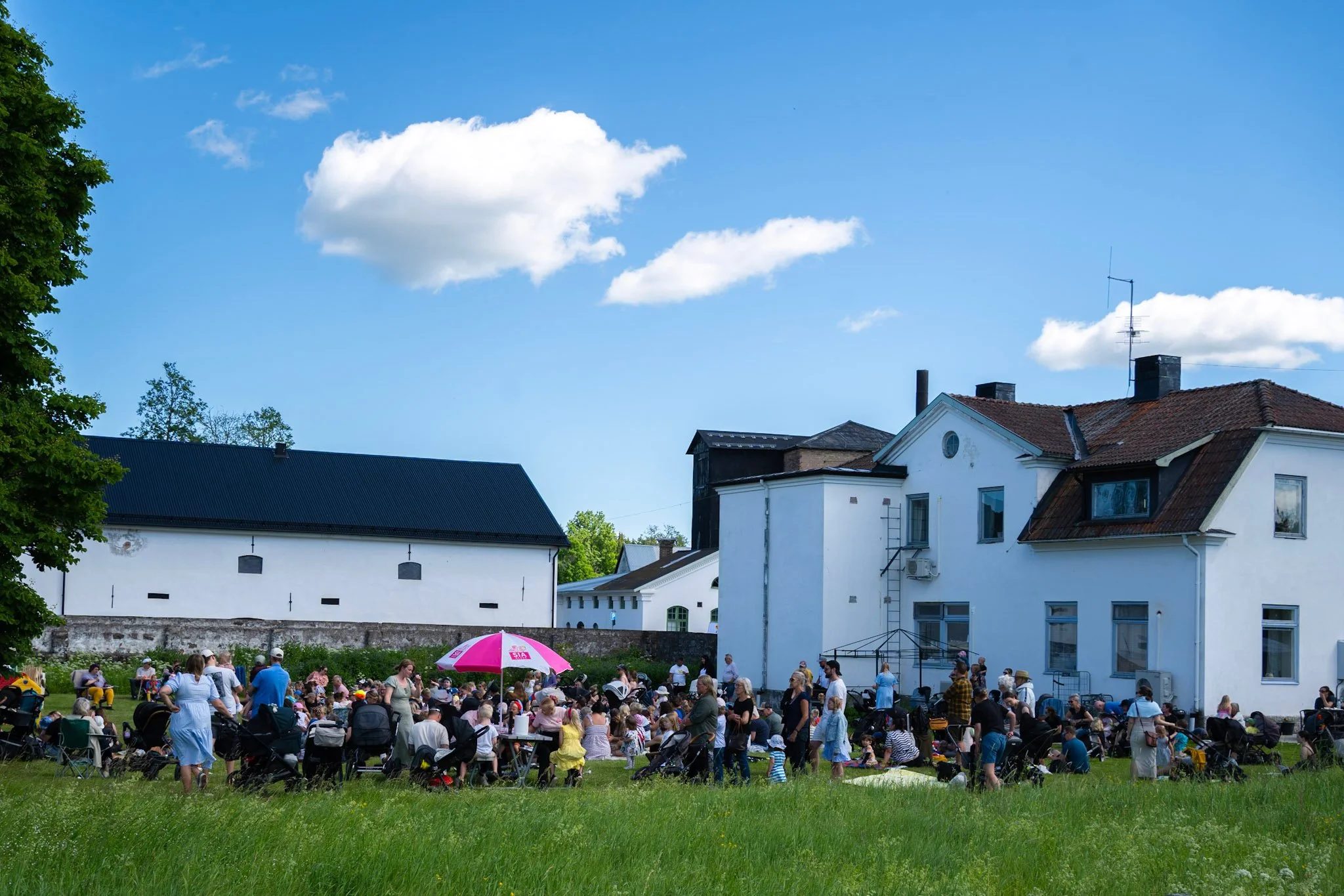 Folk samlat utomhus på gräset med solstolar och paraply, omringade av vita gamla byggnader under en blå himmel med ett par moln.