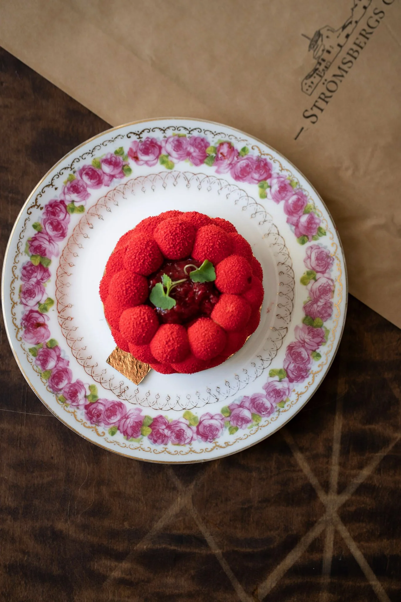 En liten röd dessert med hallon och gröna blad på ett fat med rosmönster, på ett träbord.