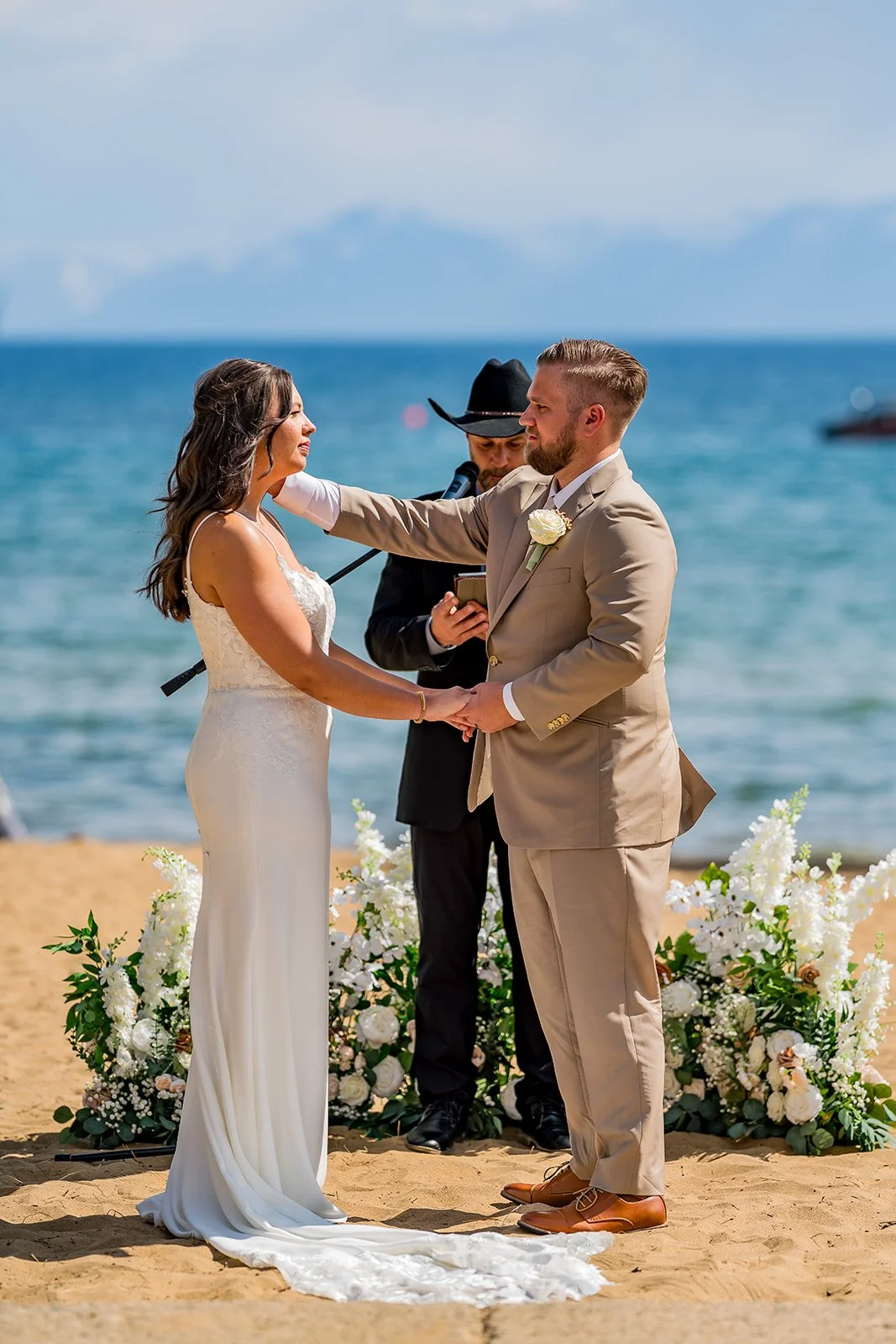  A groom in a tan suit touches his bride’s face with Lake Tahoe in the background at the North Tahoe Event Center in Kings  Beach, California. 