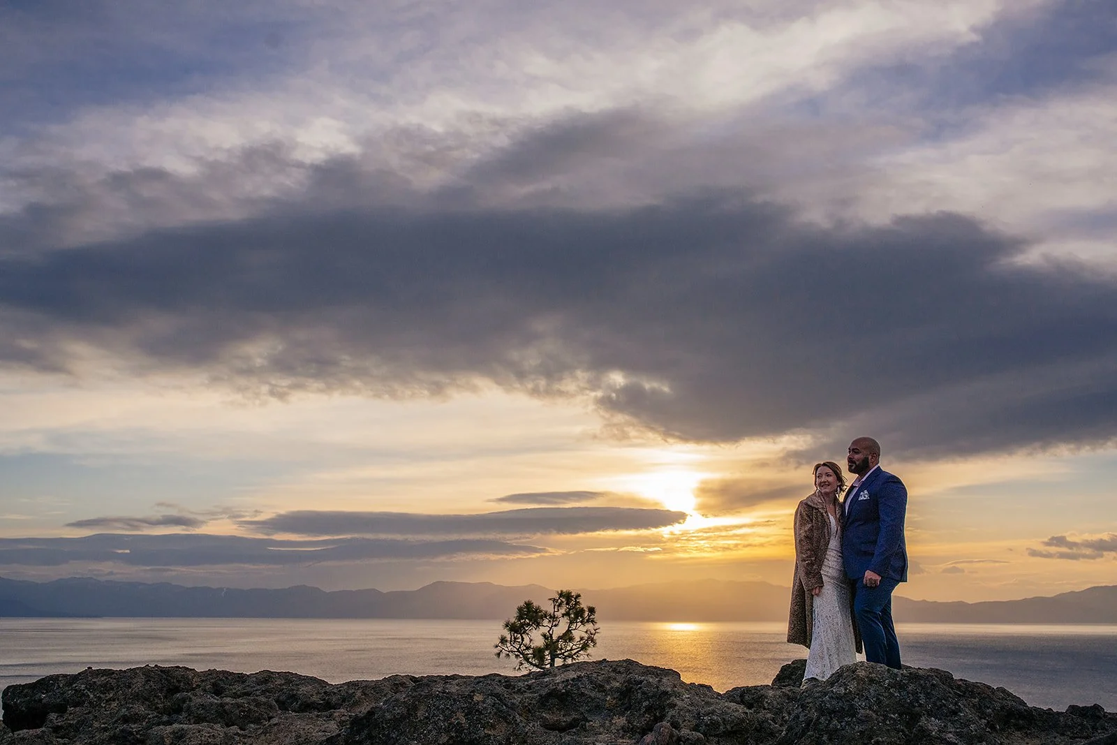 A bride wearing a fur coat and white dress stands with a groom wearing a blue suit atop Eagle Rock at Lake Tahoe with the sun rising behind them photographed by Lake Tahoe wedding photographer Nicky Byrnes Photography