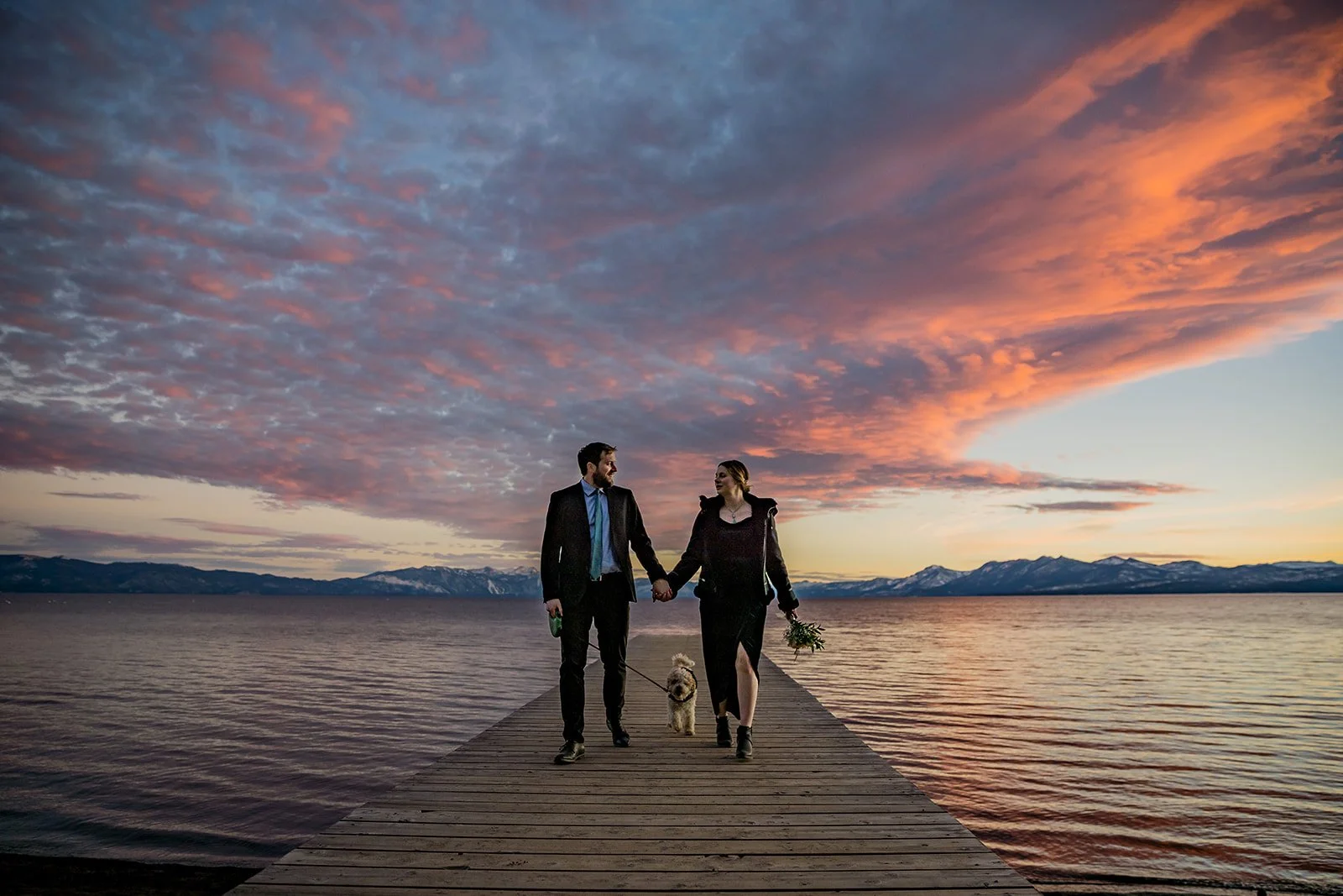 A man wearing a suit and  a woman wearing a jacket and dark dress hold hands while their small dog walks behind them at Lake Tahoe with a colorful sunset sky photographed by Lake Tahoe wedding photographer Nicky Byrnes Photography