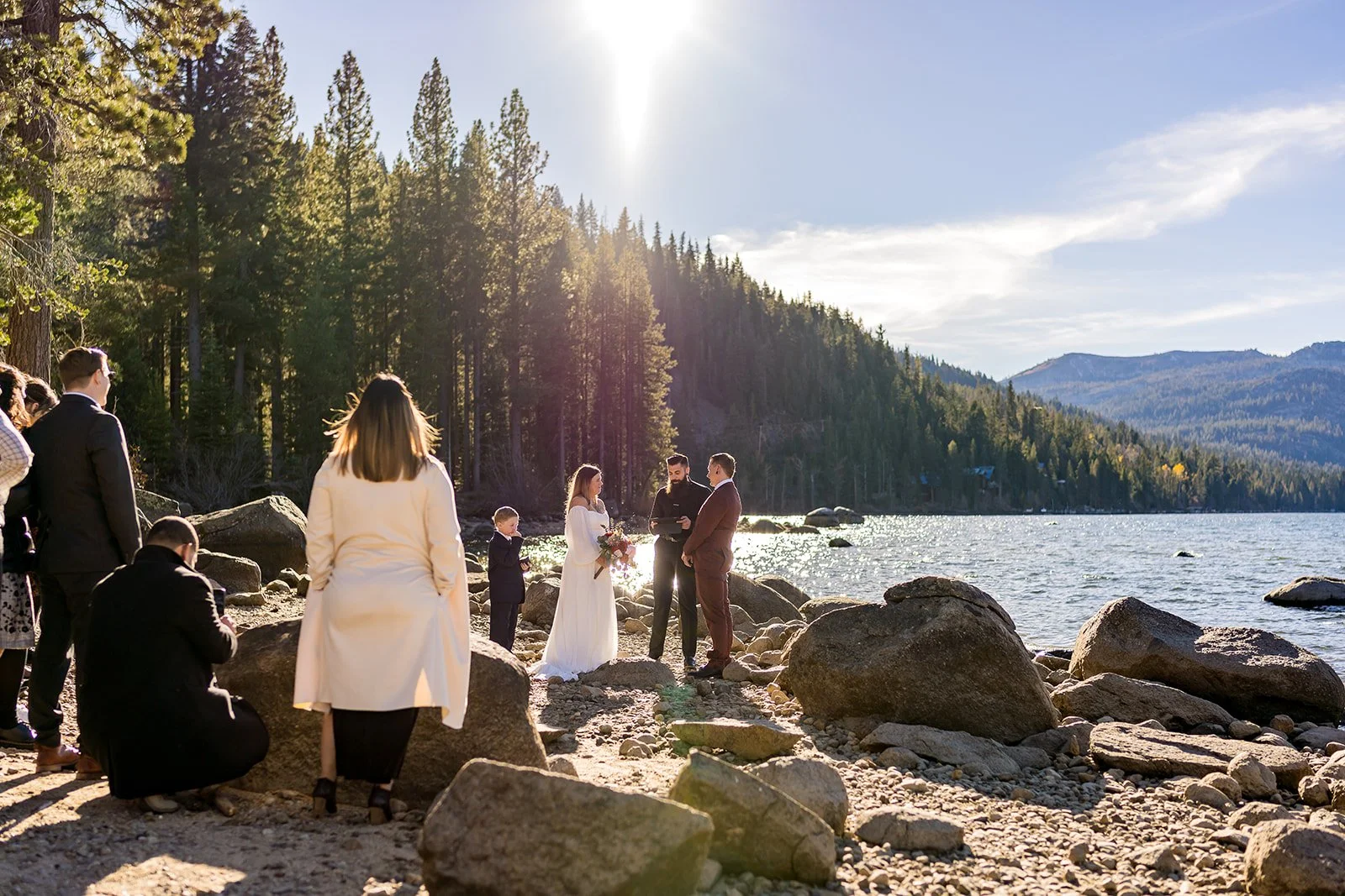 A bride and groom are surrounded by family at Donner Memorial State Park with Donner Lake and mountains in the background photographed by Lake Tahoe wedding photographer Nicky Byrnes Photography