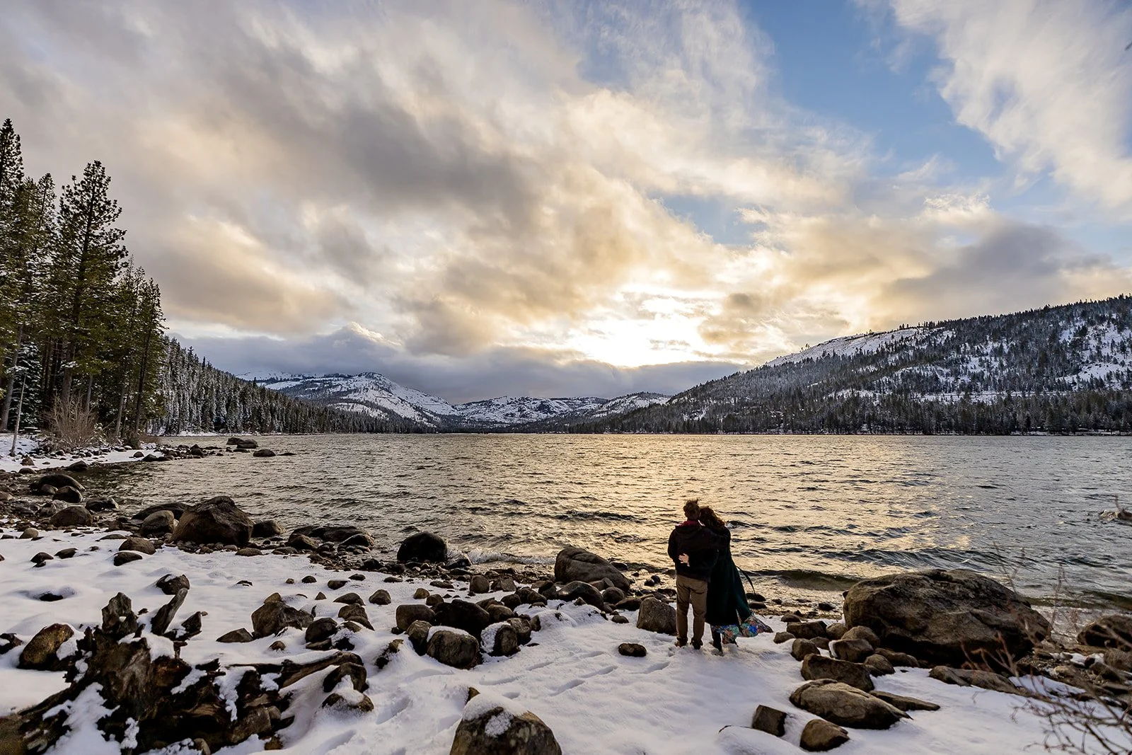 A man and a woman embrace in a snow covered landscape at Donner Memorial State Park with snow-covered mountains and Donner Lake in the background photographed by Lake Tahoe Wedding Photographer Nicky Byrnes Photography