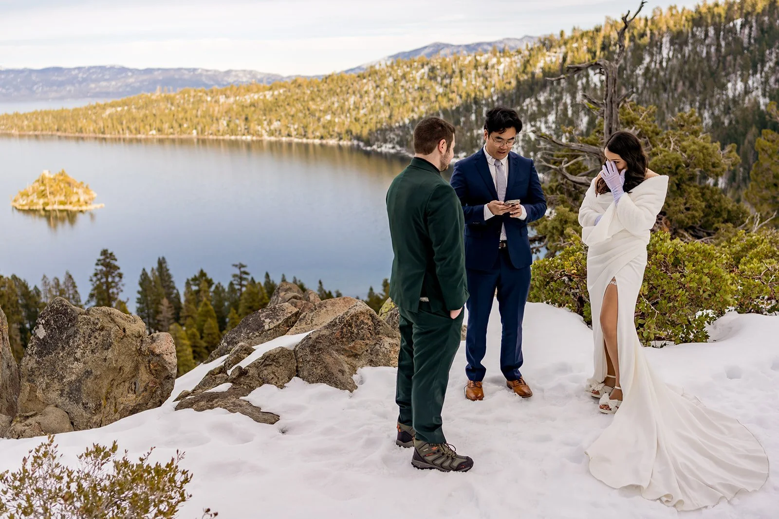 A bride wipes a tear while a groom and officiant stand in front of Emerald Bay in the winter with snow on the landscape photographed by Lake Tahoe wedding photographer Nicky Byrnes Photography