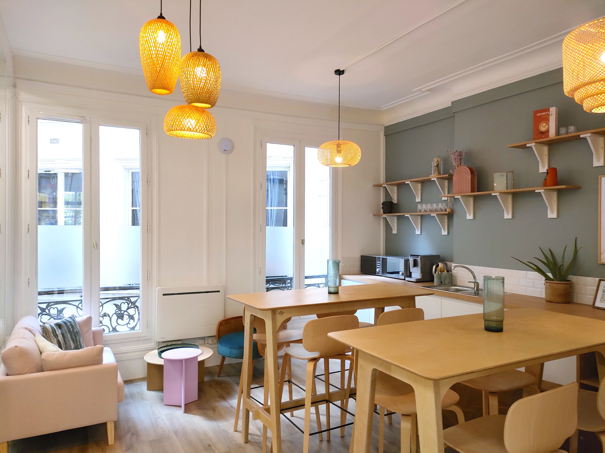 Intérieur d'une cuisine ouverte avec un salon. Éclairage avec quatre suspensions en rotin au plafond. Murs blancs et mur vert pastel avec étagères en bois. Plusieurs fenêtres laissant entrer la lumière. Mobilier en bois clair, table, chaises et étagères décoratives. Plateau de cuisine avec appareils électroménagers, plantes et objets décoratifs.