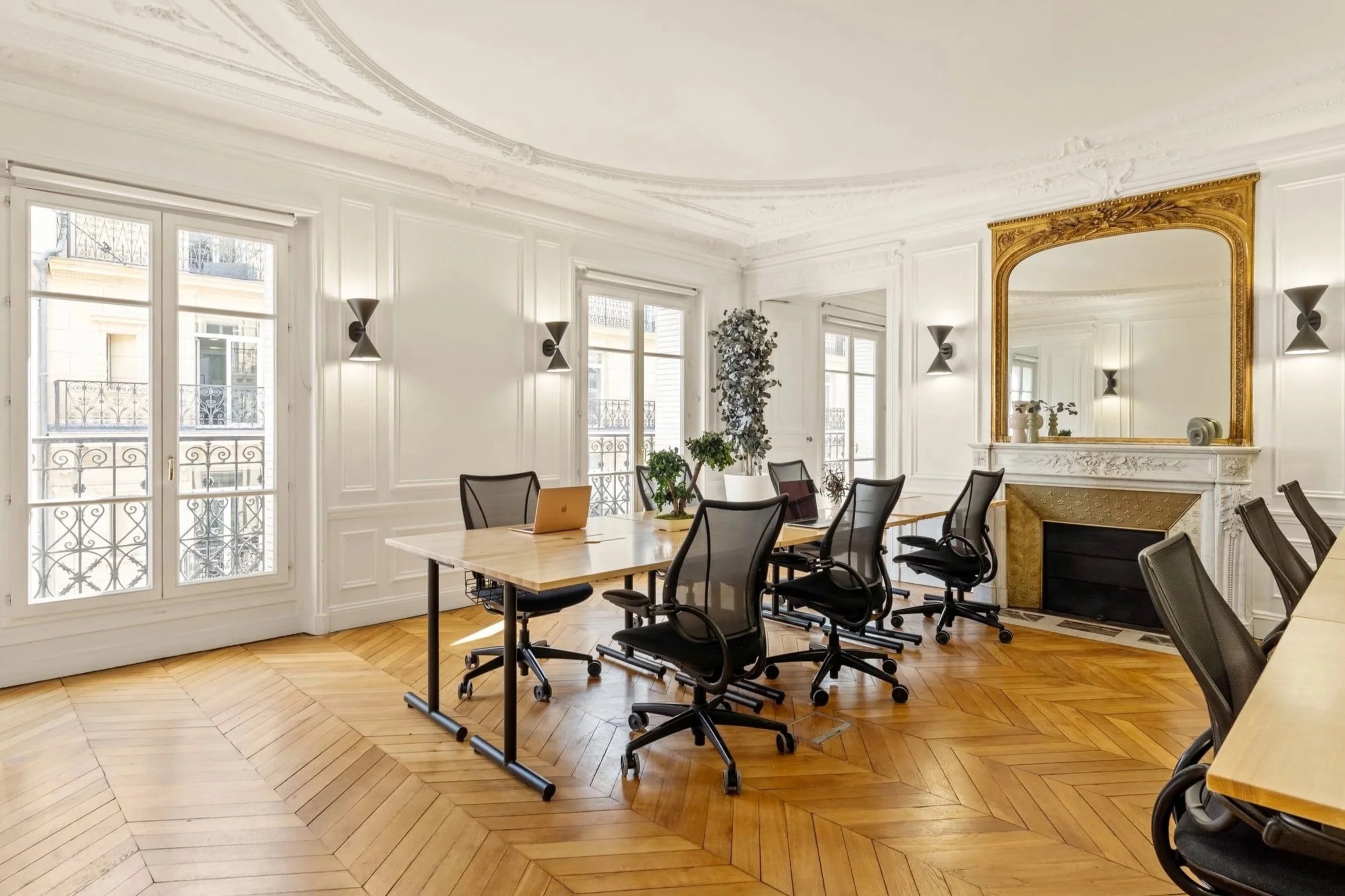 Salle de réunion élégante avec fenêtres, miroir ornée de dorures, cheminée en marbre, fauteuils de bureau et table en bois clair