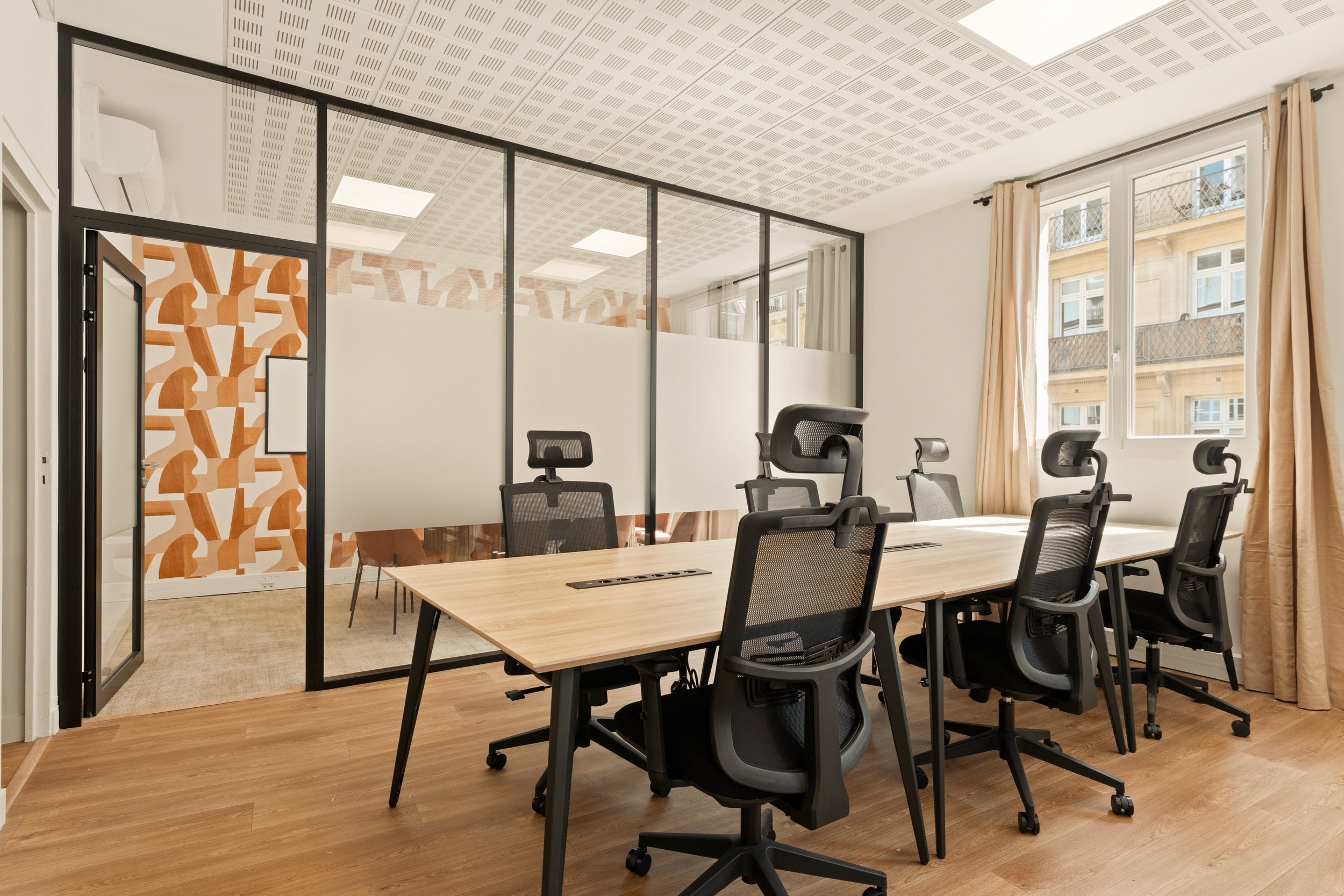 Salle de réunion moderne avec une grande table en bois entourée de chaises noires, séparée par une cloison en verre avec un motif géométrique, grande fenêtre laissant entrer la lumière naturelle.