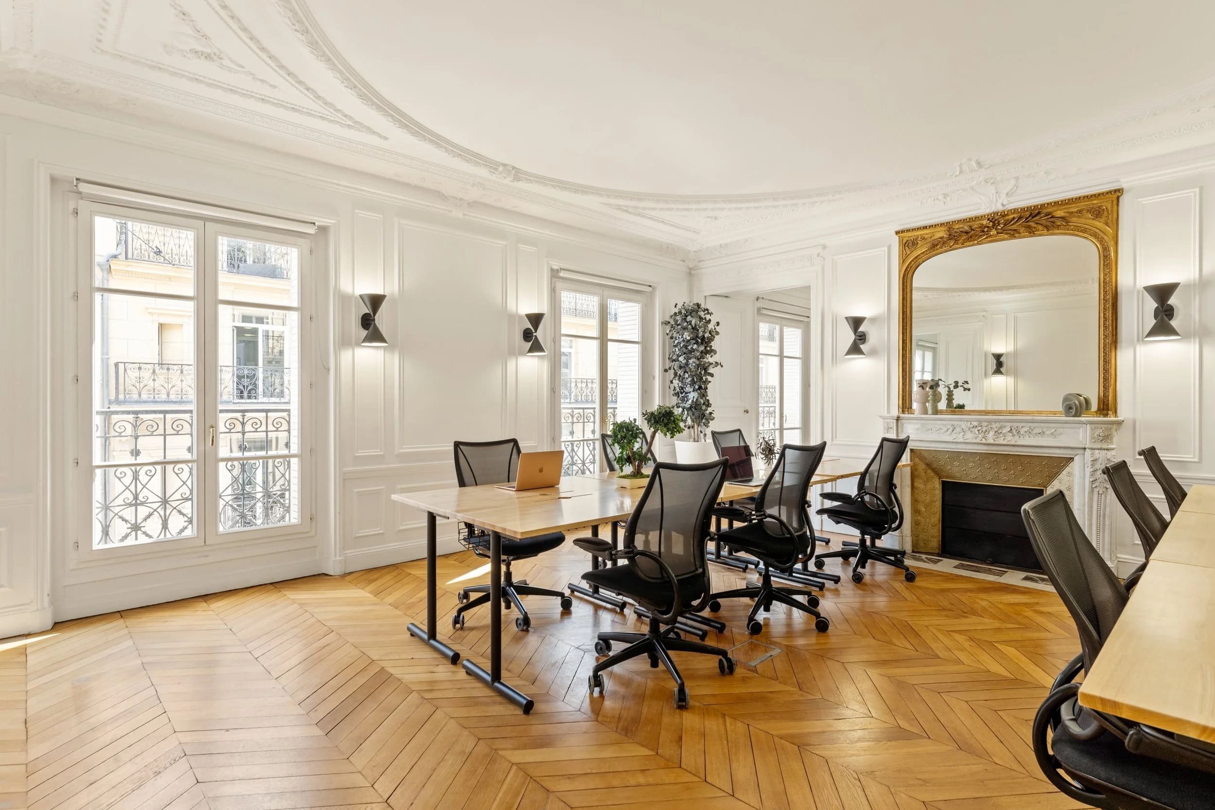 Salle de réunion élégante avec grand miroir doré au-dessus de la cheminée, fenêtres avec vues urbaines, chaises de bureau et table en bois clair.
