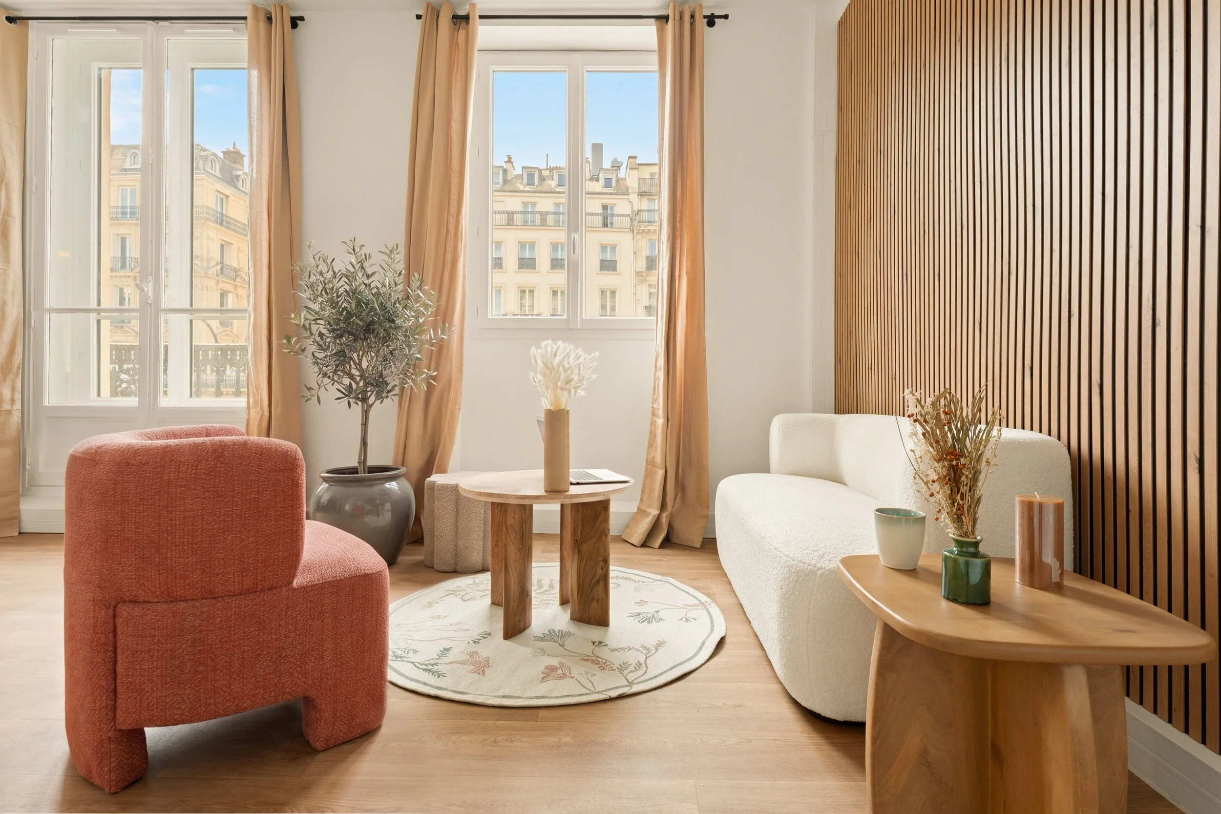 Un espace détente moderne dans le bureau Grands Boulevards avec de grandes fenêtres, des rideaux beige, un canapé blanc, un fauteuil rose, plusieurs vases avec des plantes et des décorations en bois.