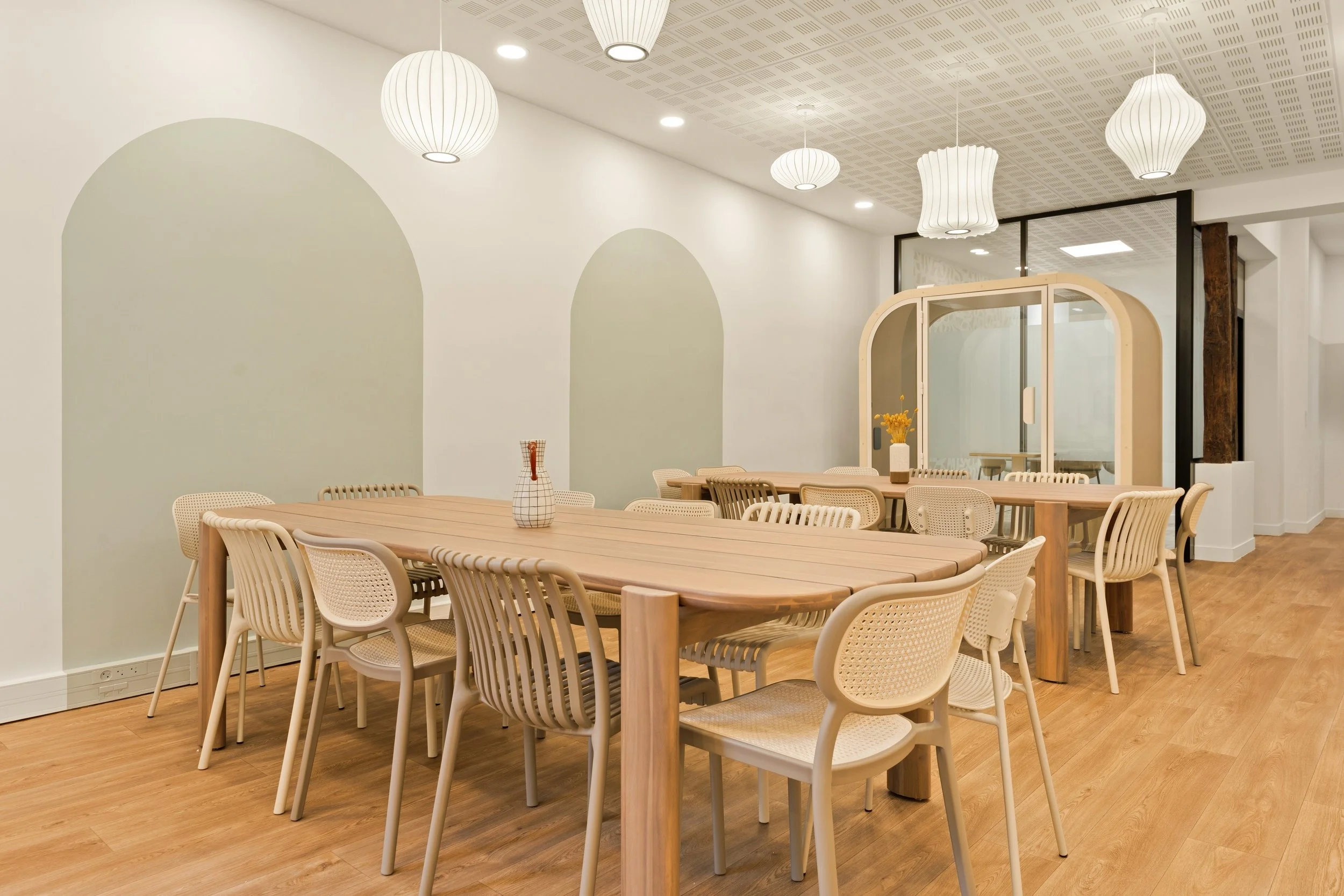 Une salle à manger moderne avec une grande table en bois clair, plusieurs chaises et bancs, murs blancs, arched design, et une lumière suspendue.
