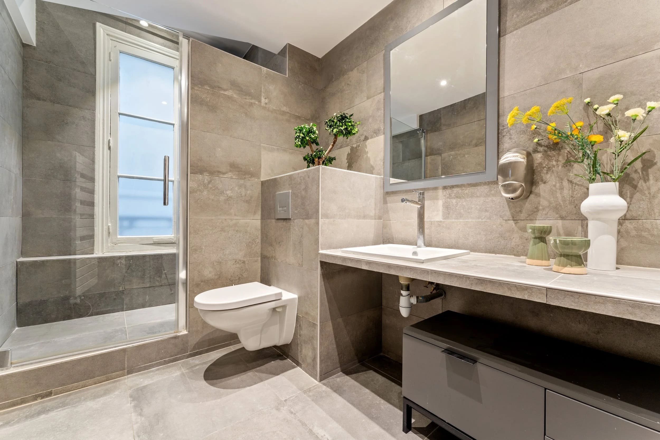 Une salle de bain moderne avec des murs et un sol en carreaux gris, un lavabo avec un miroir rectangulaire au-dessus, une plante verte dans un pot blanc, et des vases décoratifs avec des fleurs, une fenêtre avec volet en bois, et un toilette suspendu.