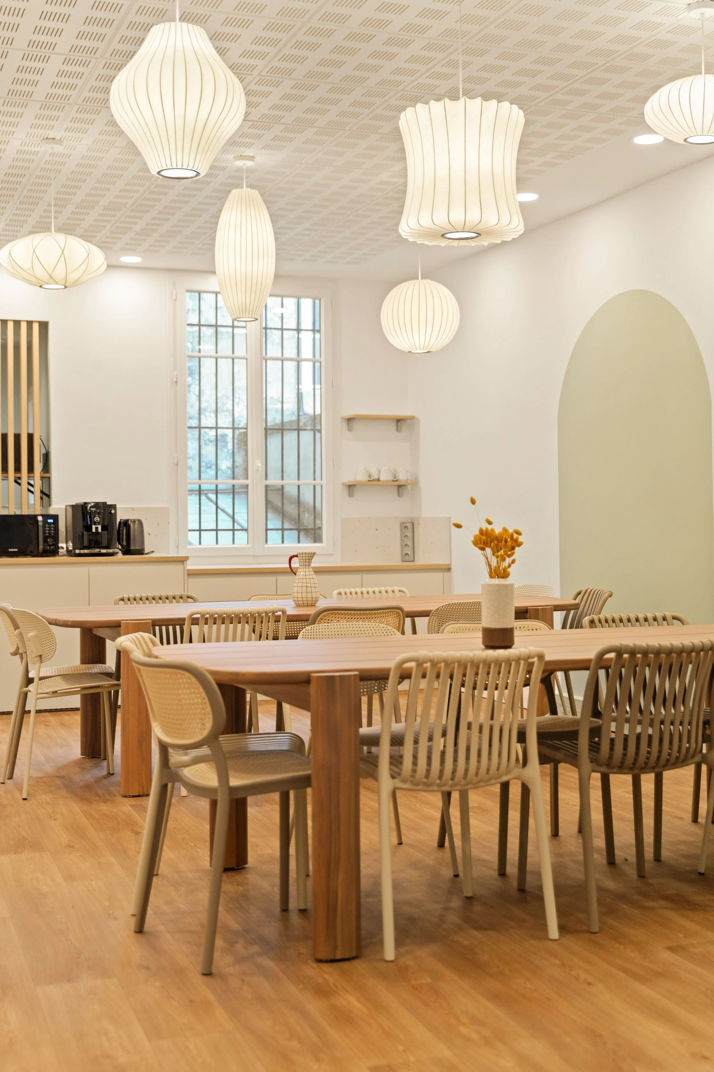 Salle à manger et cuisine moderne du bureau Grands Boulevards avec plusieurs lampes suspendues blanches, fenêtres avec grille, et mobilier en bois clair avec des chaises autour de deux grandes tables.
