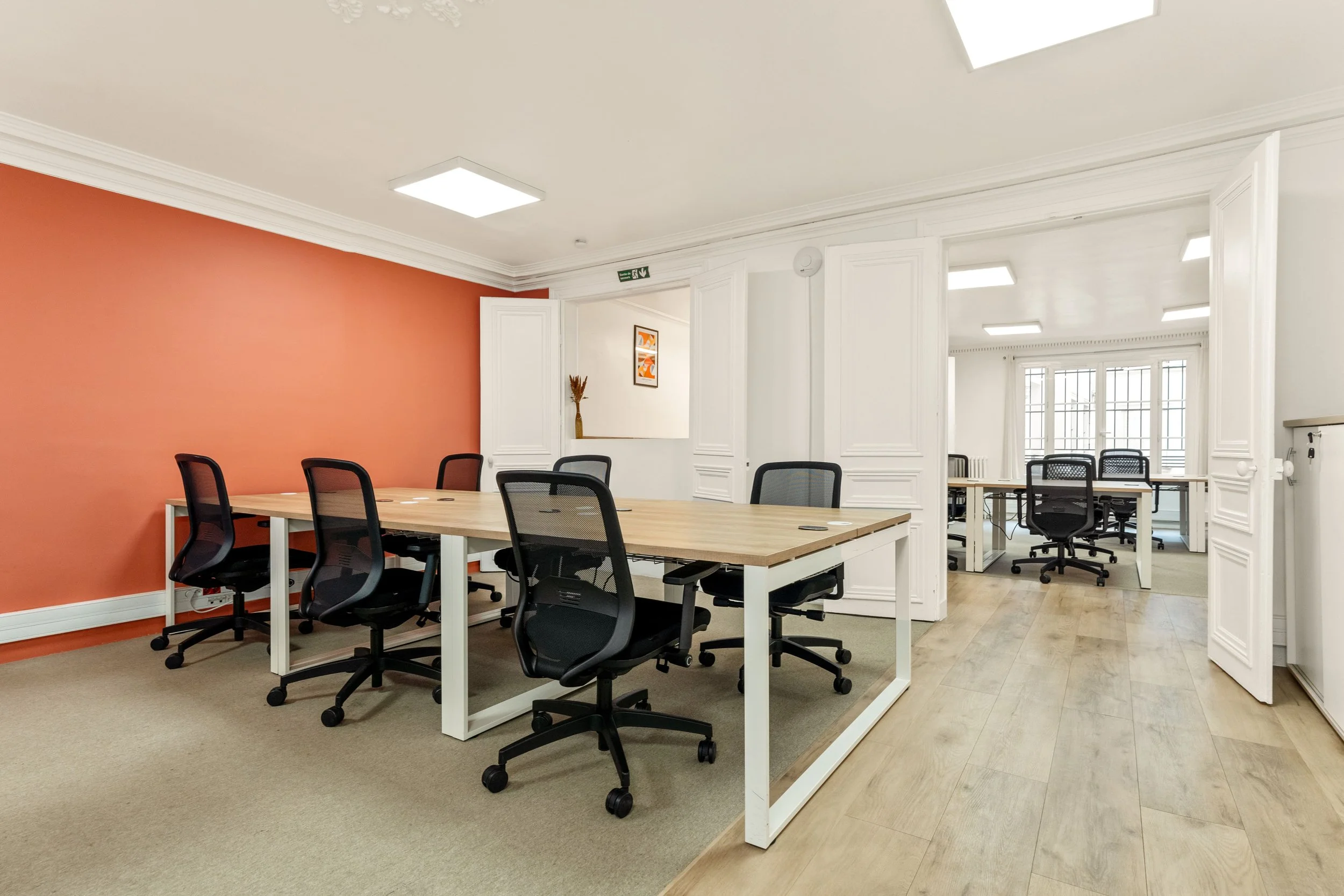 Une salle de réunion avec des tables en bois et des chaises de bureau noires. Les murs sont blancs, avec une cloison en bois clair et un mur accent en orange. La pièce est bien éclairée par des plafonniers lumineux.