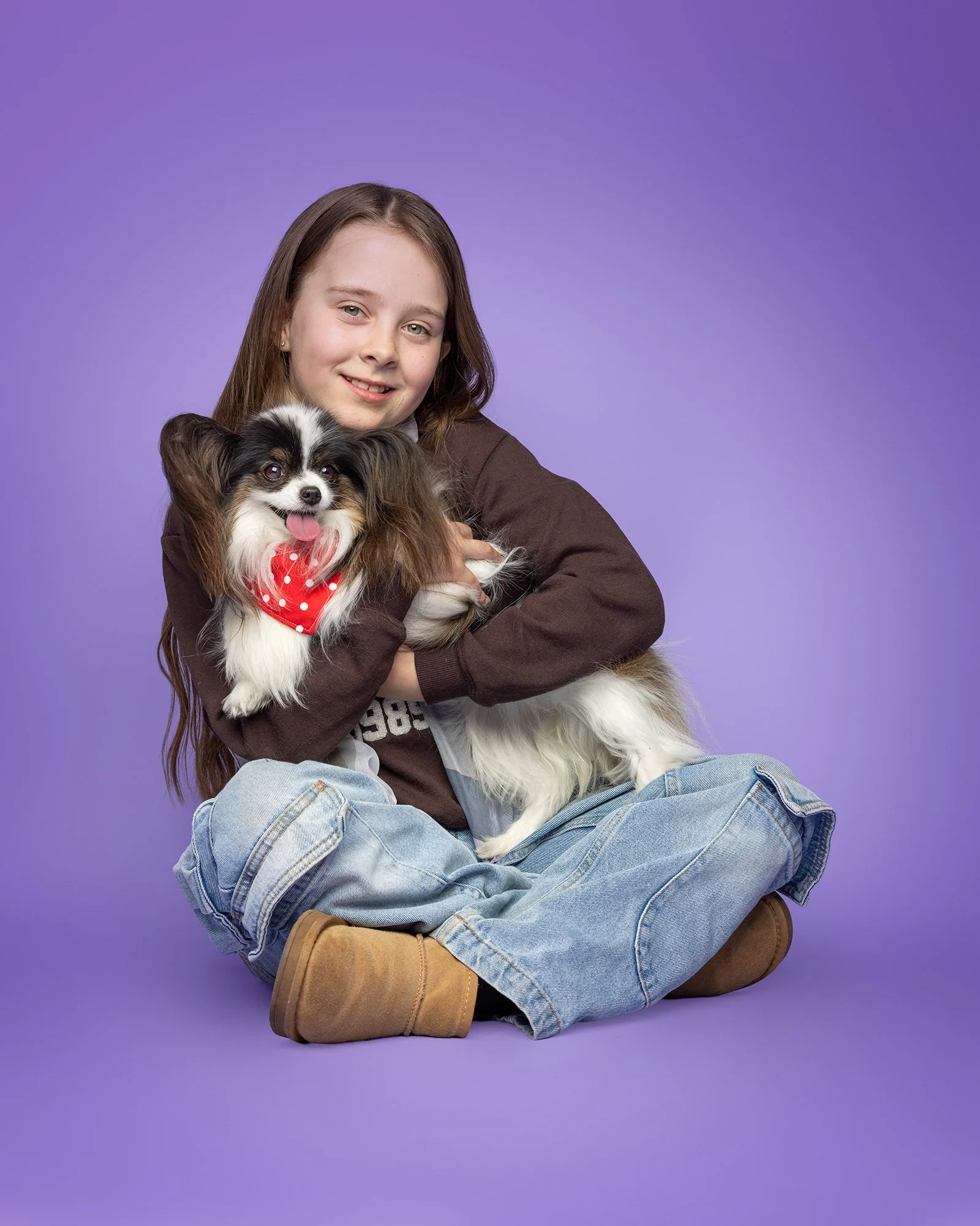 a little girl with brown hair sitting cross legged with her papillion dog on her lap on a purple studio background