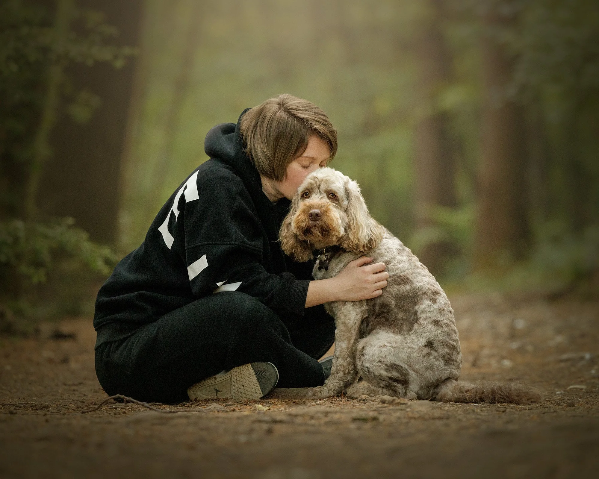 a woman sitting with her cockerpoo dog, kissing his head while he looks at. the camera in selsdon nature reserve on his dog photography session with london pet photographer amie barron