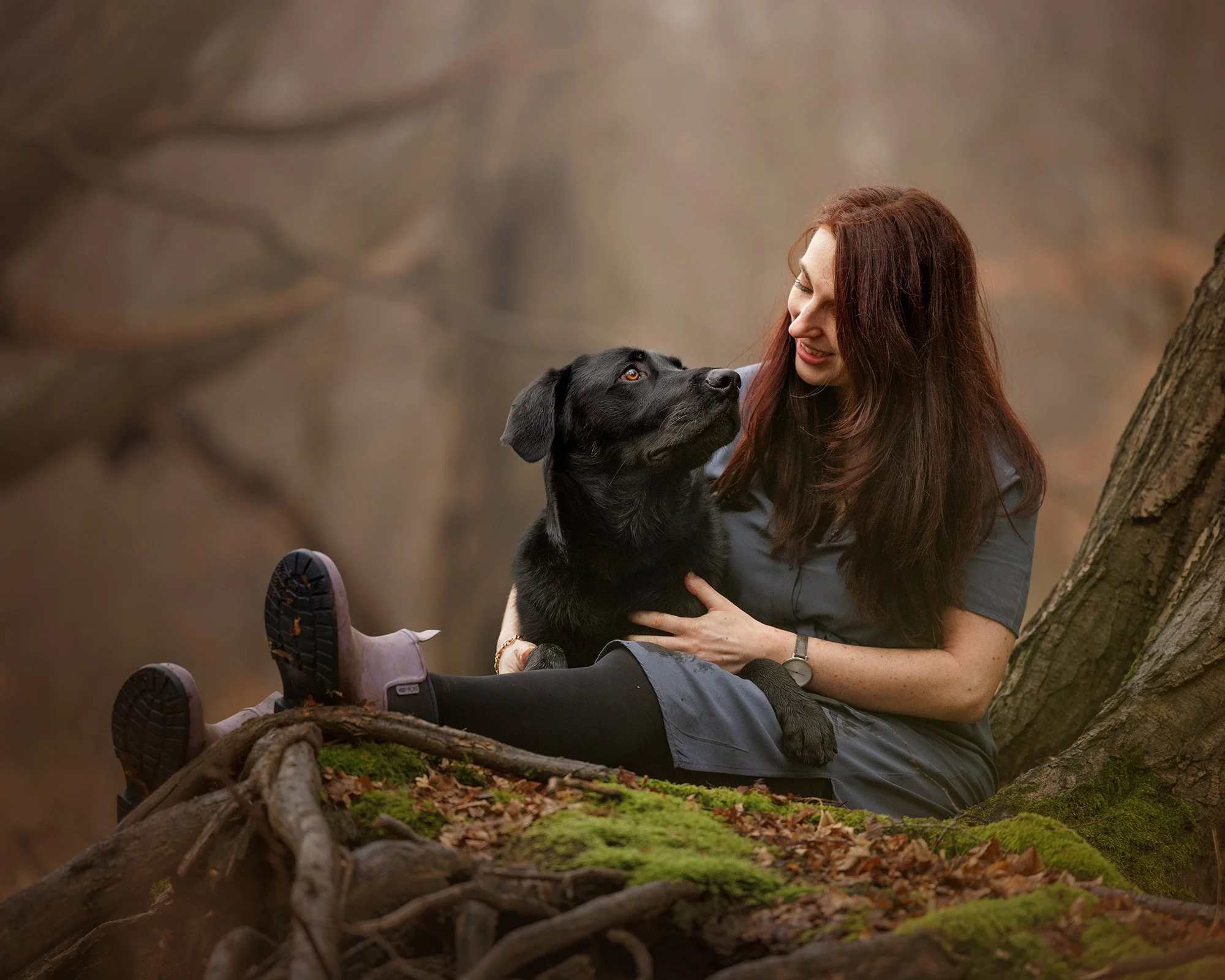 A woman with long red hair sitting in Epping forest, holding a black Labrador retriever with brown eyes, both smiling and looking at each other.