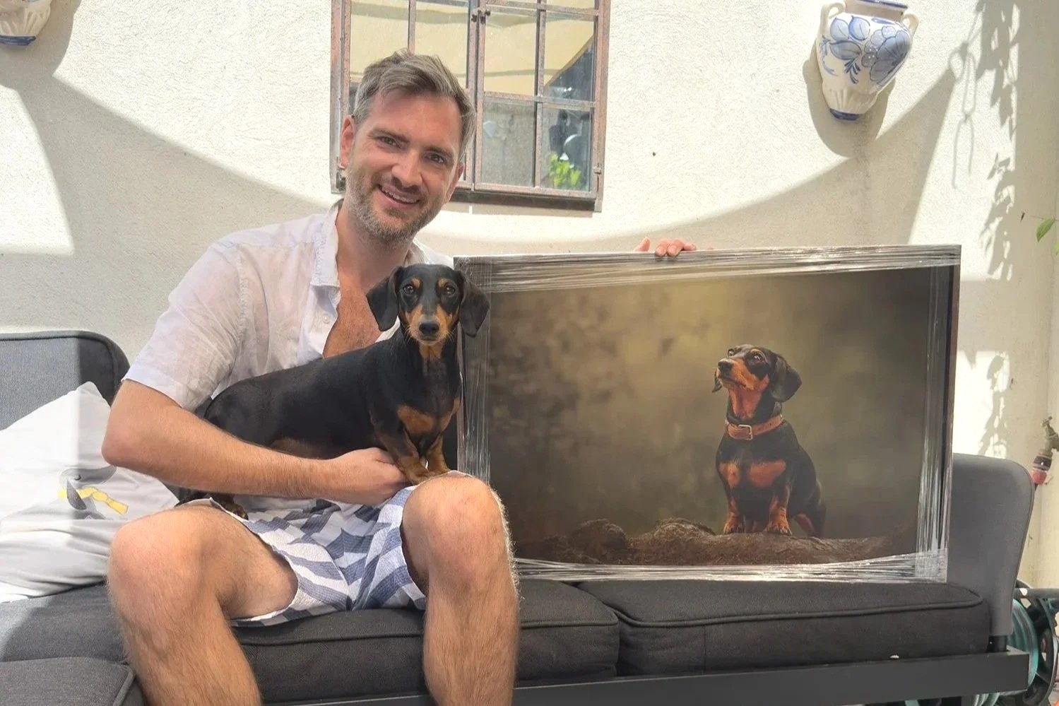 a man sitting with his black and tan dachshund on his lap holding a large piece of wall art of the same dachshund dog