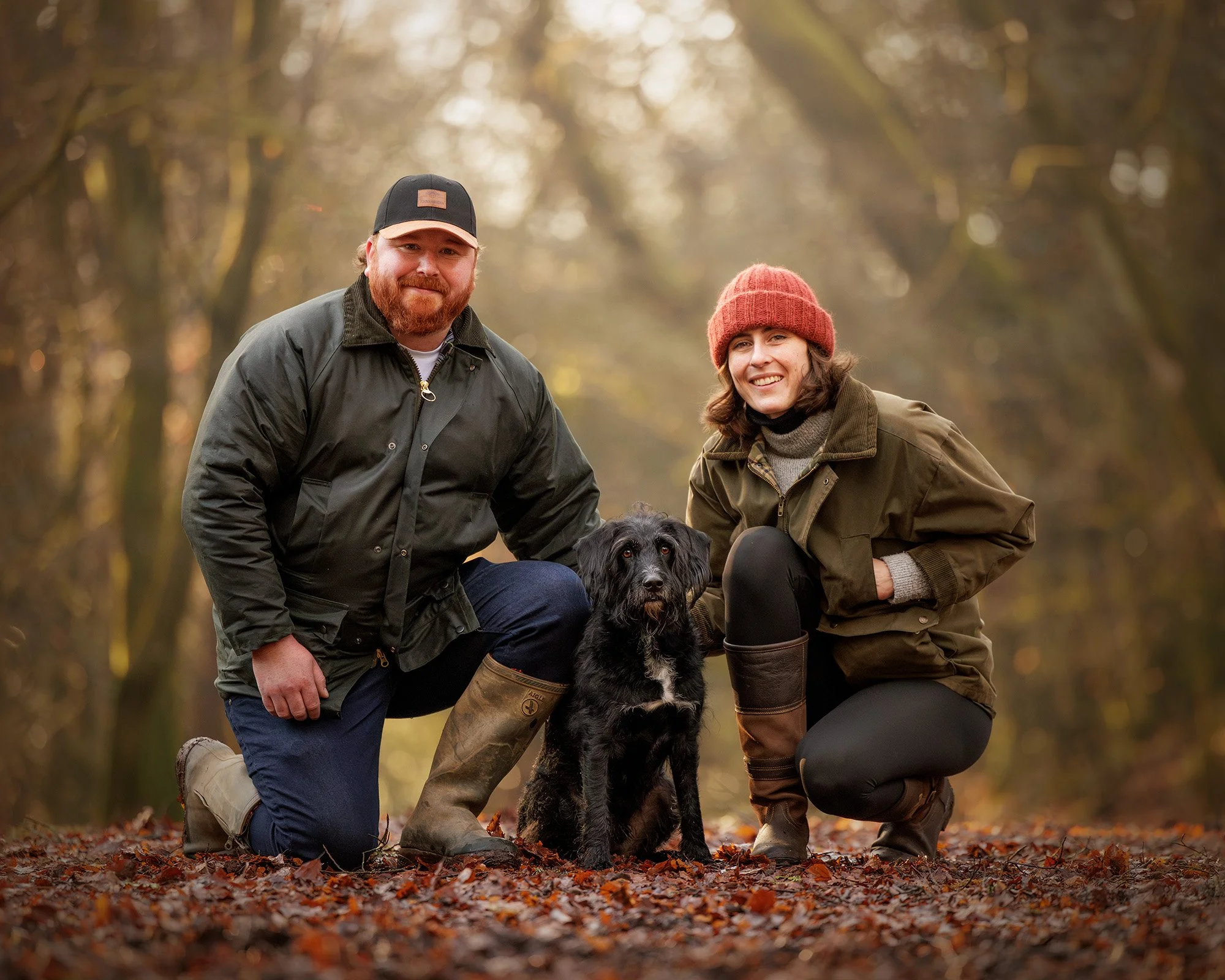 A man and woman outdoors in a wooded area during fall, kneeling on fallen leaves with a black and white dog between them. The man has a beard, wears a black cap, and a dark jacket. The woman has curly hair, wears a red knit hat, a green jacket, and b