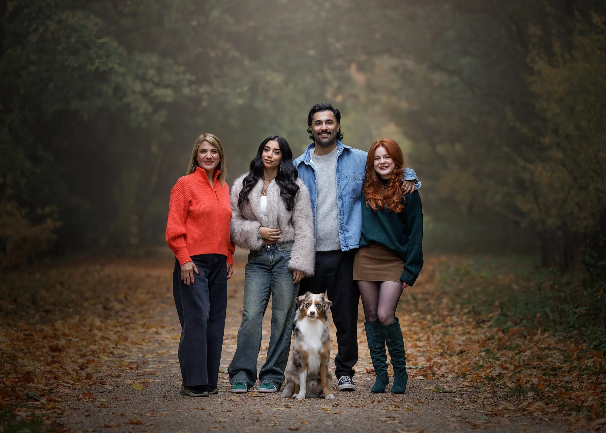 Group of five people and a dog standing on a leaf-covered path in a wooded area during autumn. Taken by London dog photographer Amie Barron in Lee Valley Park