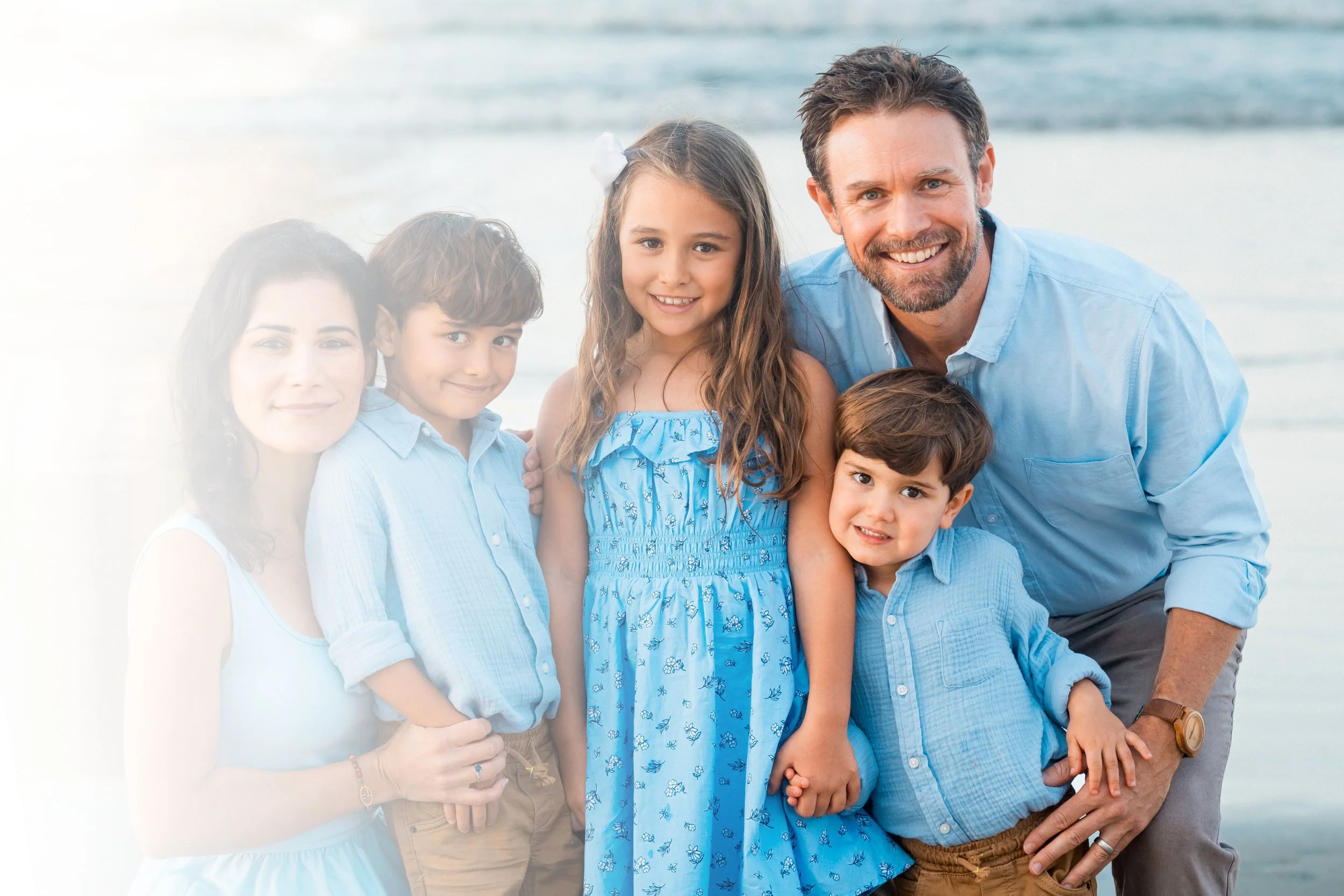A family of three, with their dog, standing on the beach at sunset.