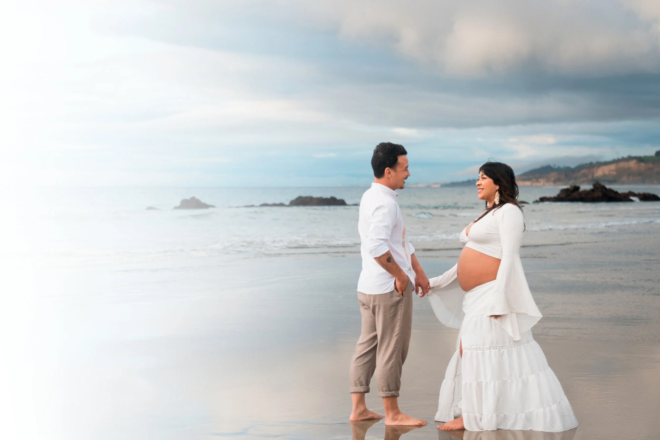 A pregnant woman in a white dress holding hands with a man on a beach with waves and rocky cliffs in the background.