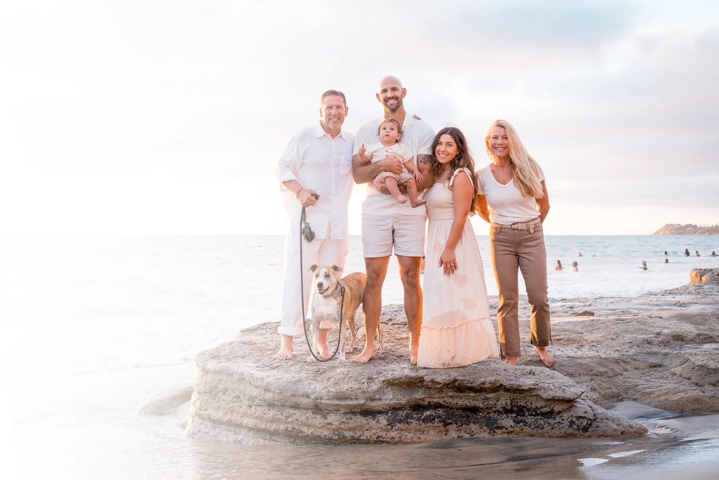 A family of five with a dog standing on a large rock on the beach at sunset, smiling at the camera.