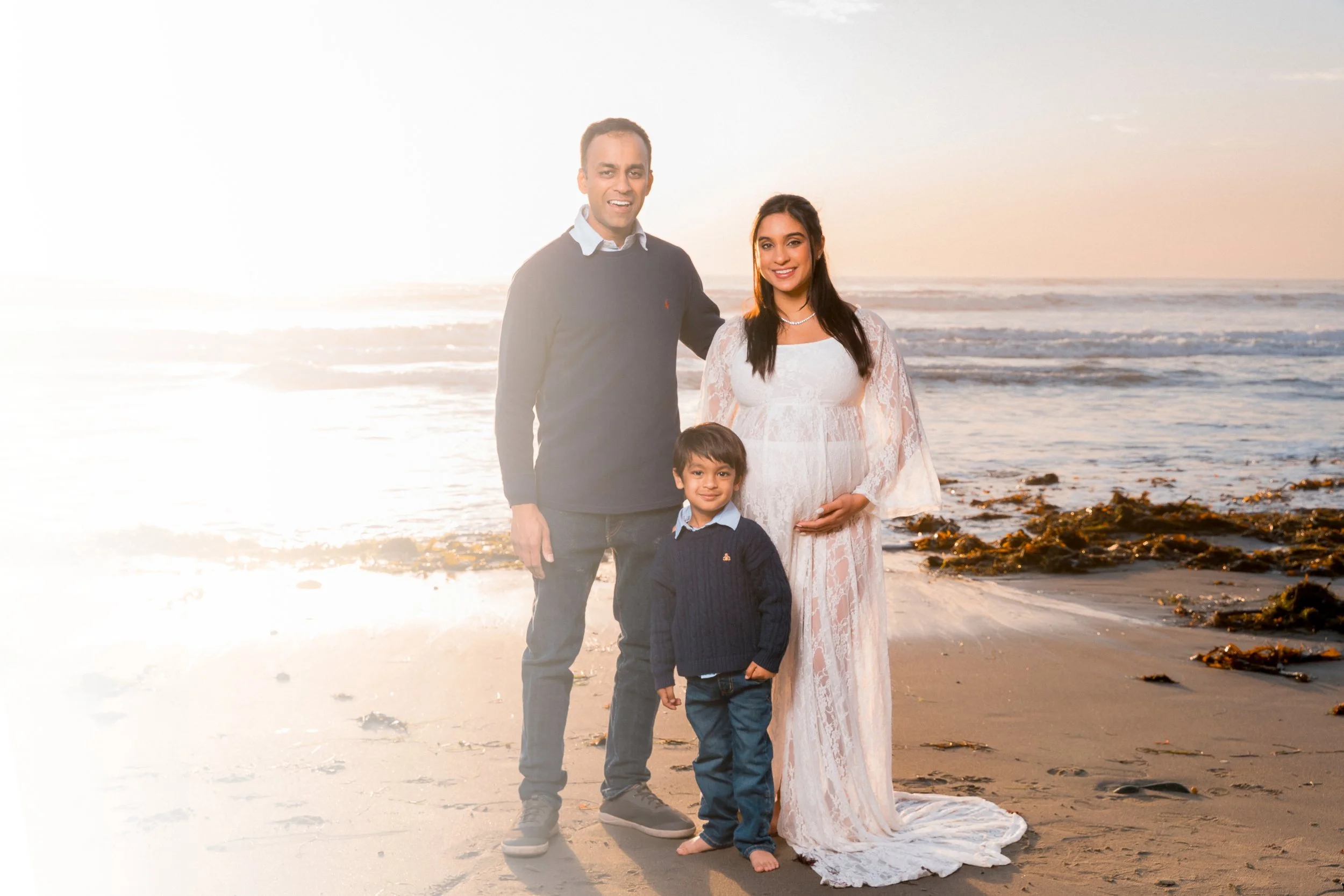 A family of three standing on a beach at sunset. The pregnant woman is in a white lace dress, the man and boy are in dark sweaters. The woman is holding her belly, the boy is smiling, and the man has his arm around the woman. The ocean and sky are in the background.