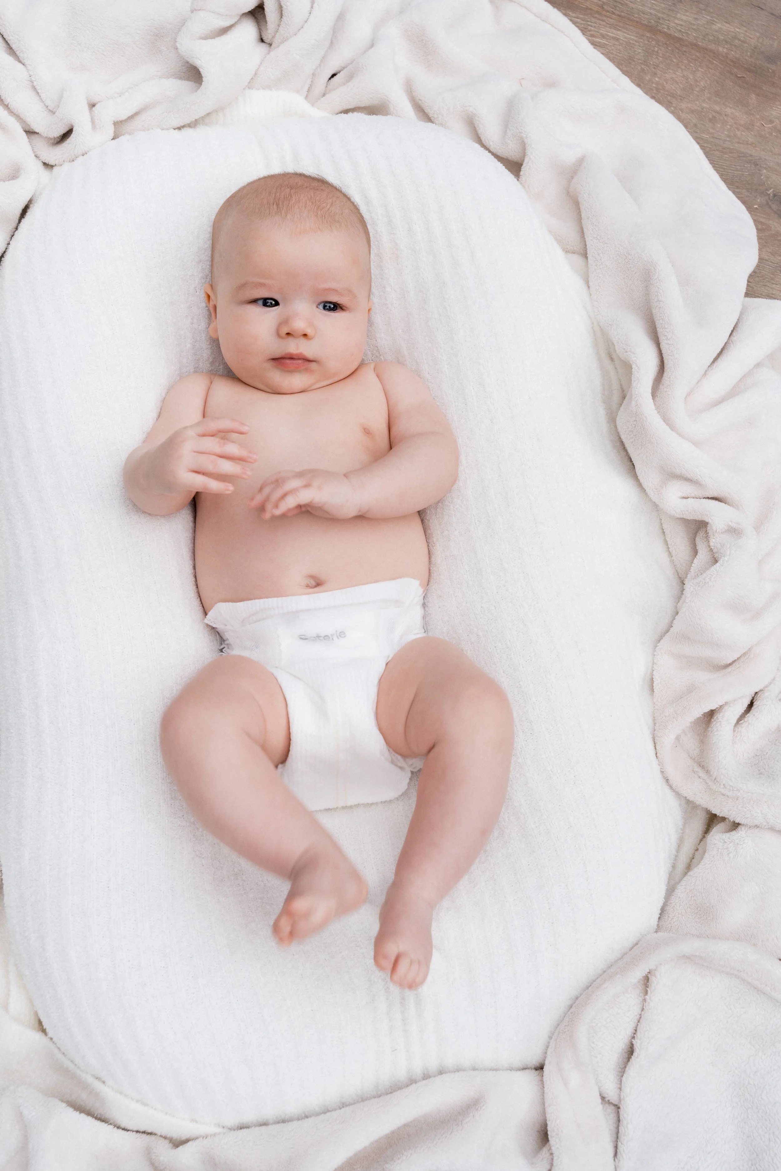 A baby lying on a white padded surface surrounded by cream-colored blankets.