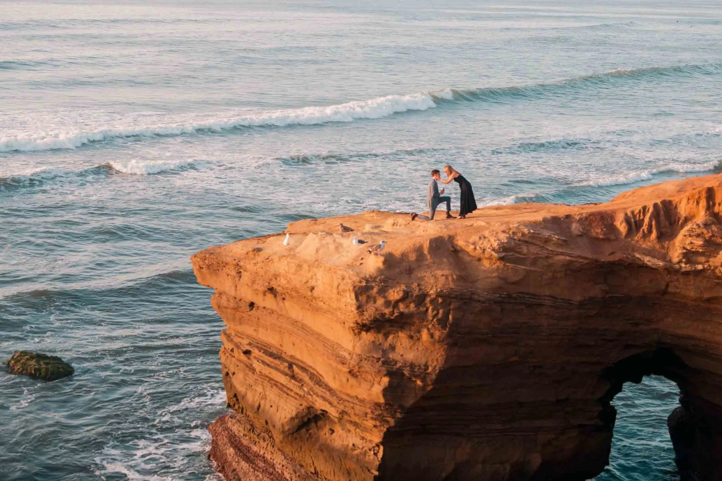 A couple on a rocky cliff by the ocean, with one person kneeling and proposing to the other. The ocean waves can be seen in the background.