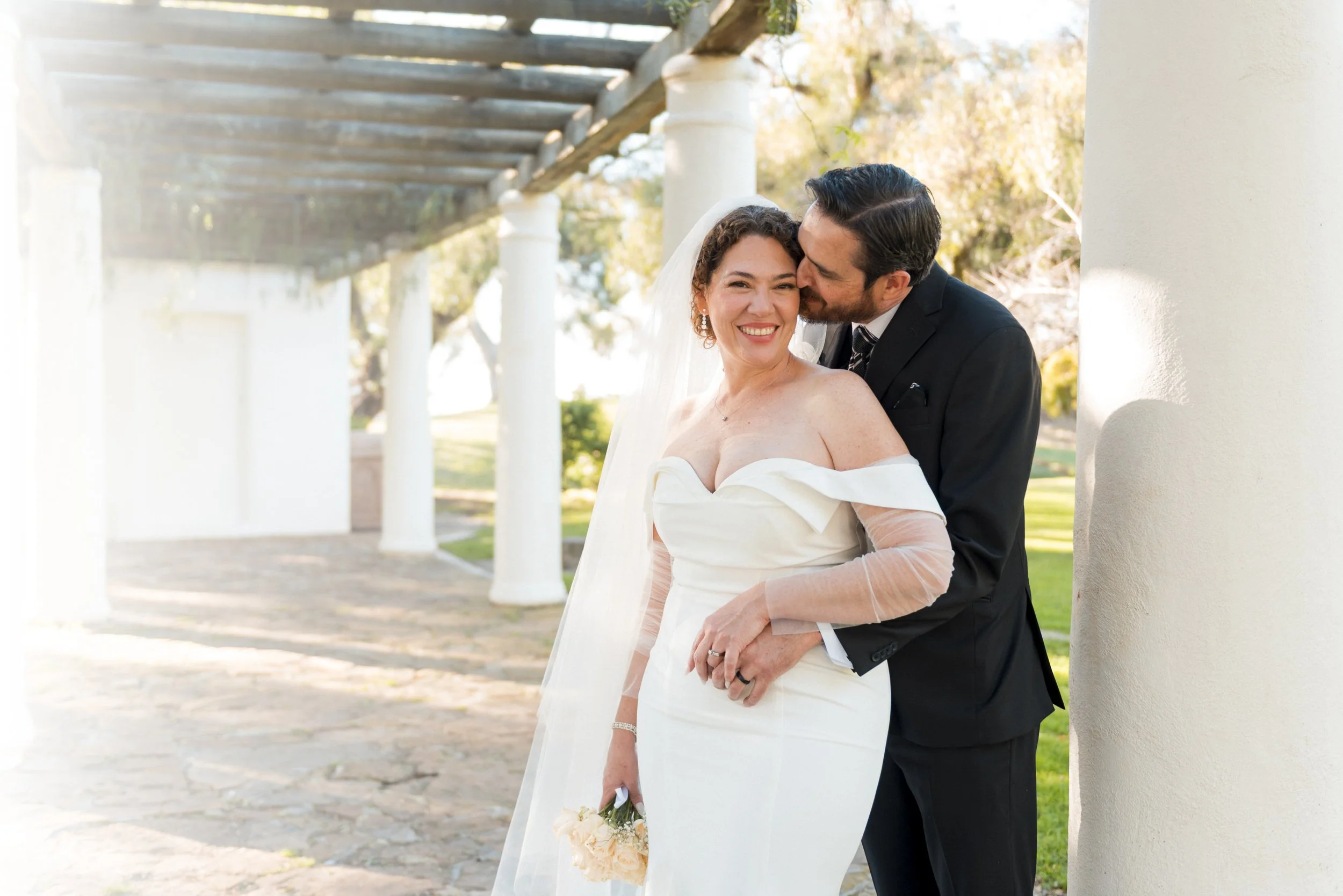 A bride and groom kissing on a beach during sunset, with rocks in the background and the ocean waves behind them.
