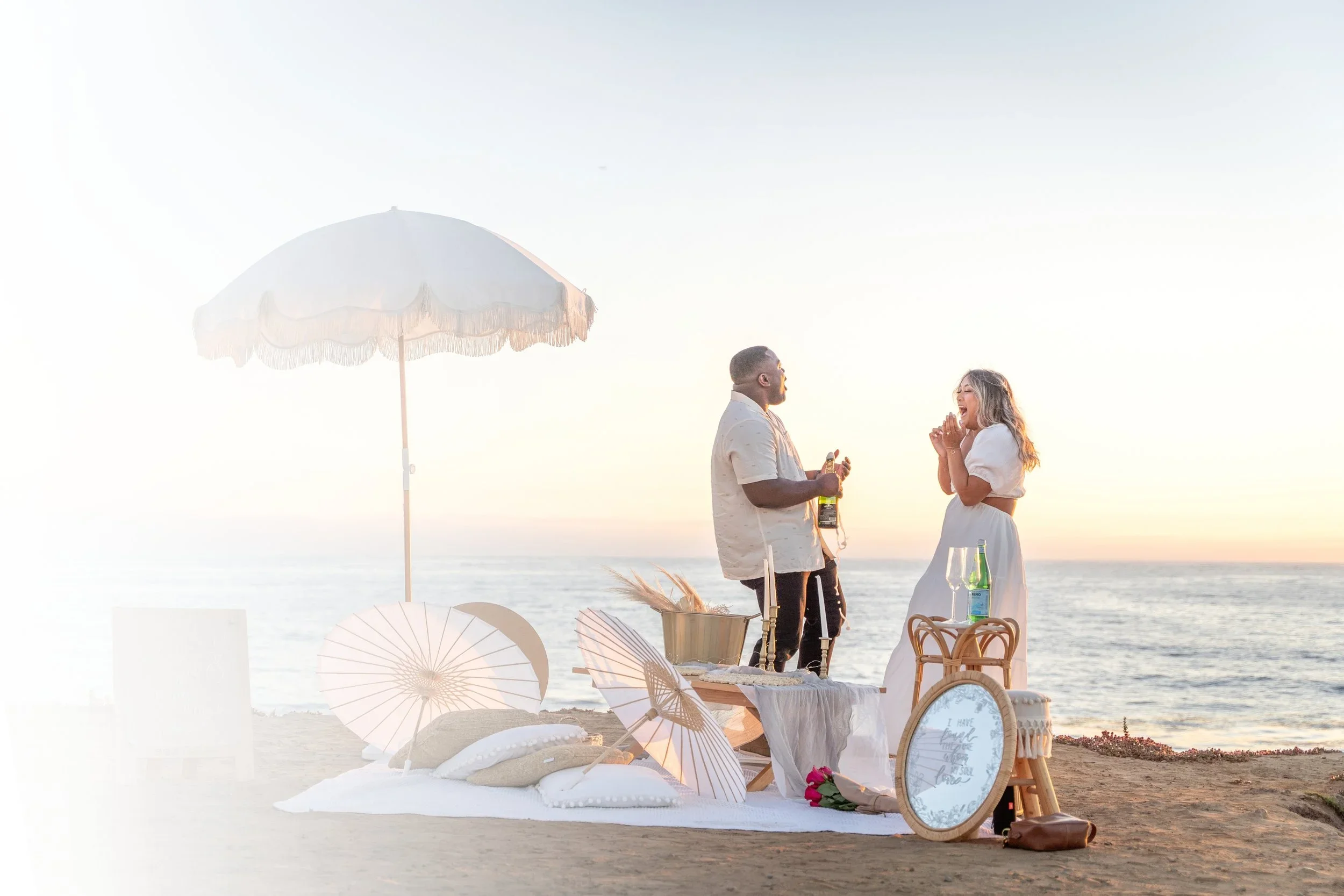 A couple on the beach having a romantic moment during sunset, with parasols, pillows, and a white blanket around them.