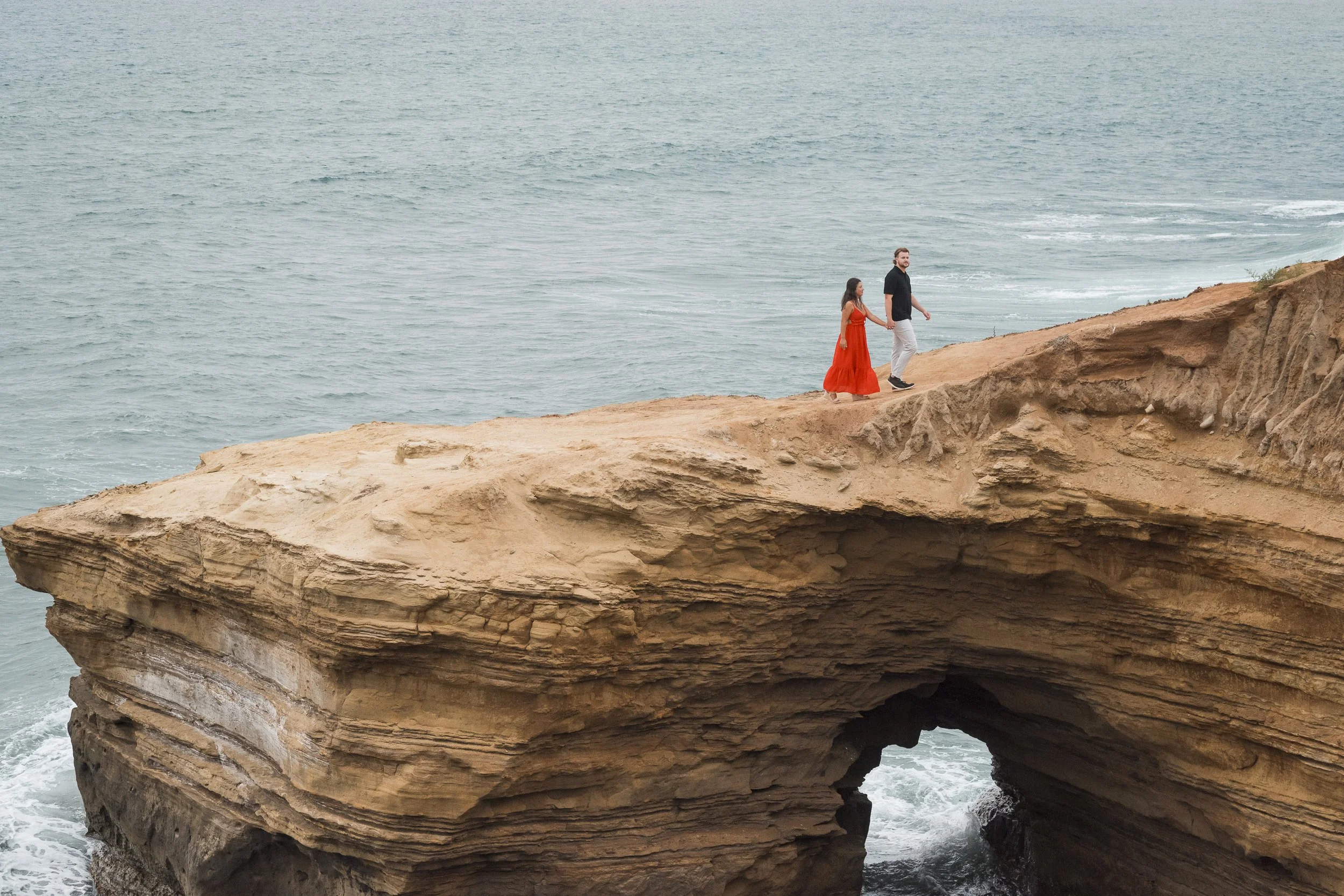 A couple holding hands walking along the edge of a large rocky cliff overlooking the ocean.