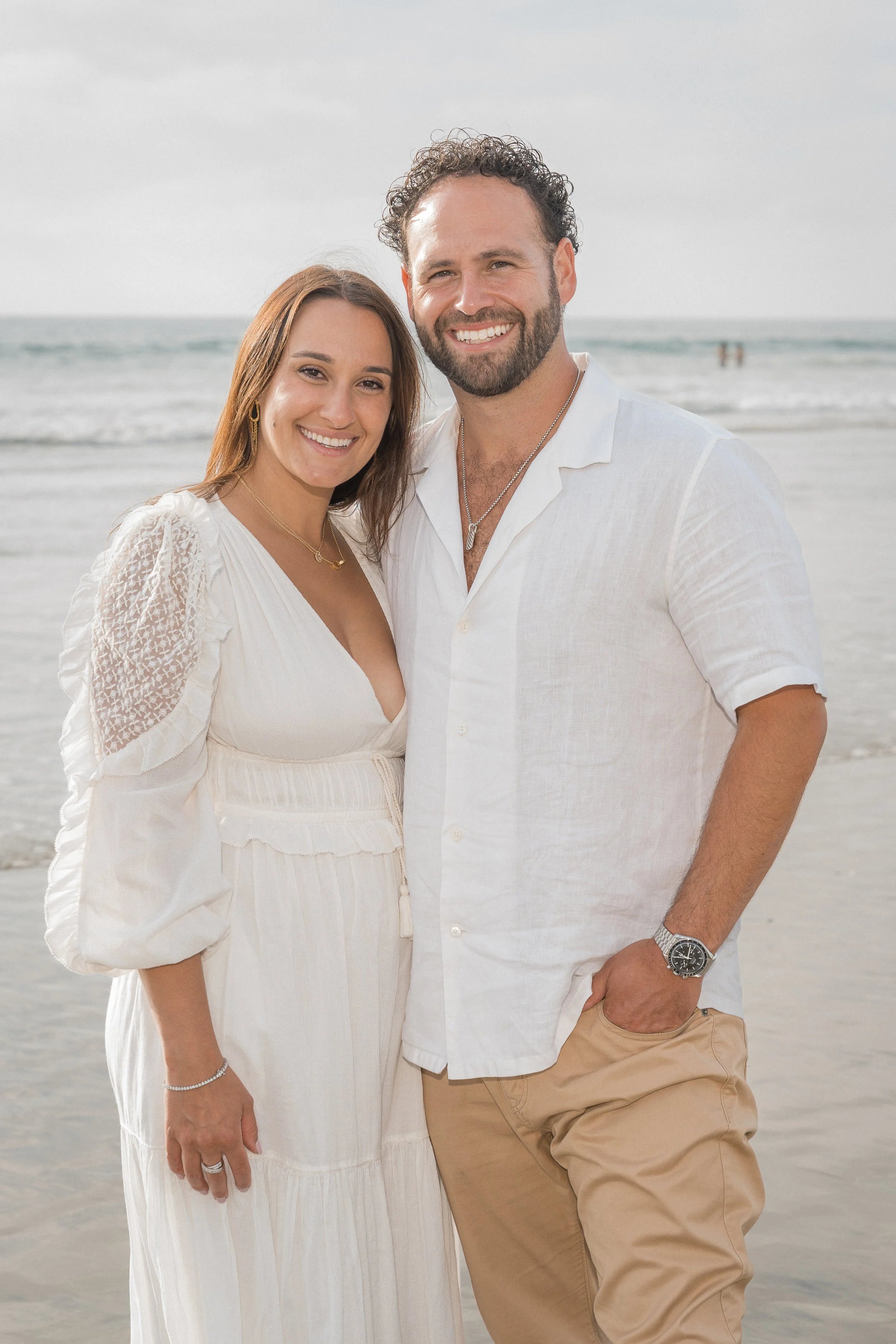 A family of three standing on the beach at sunset, smiling, with a rock formation and ocean in the background.