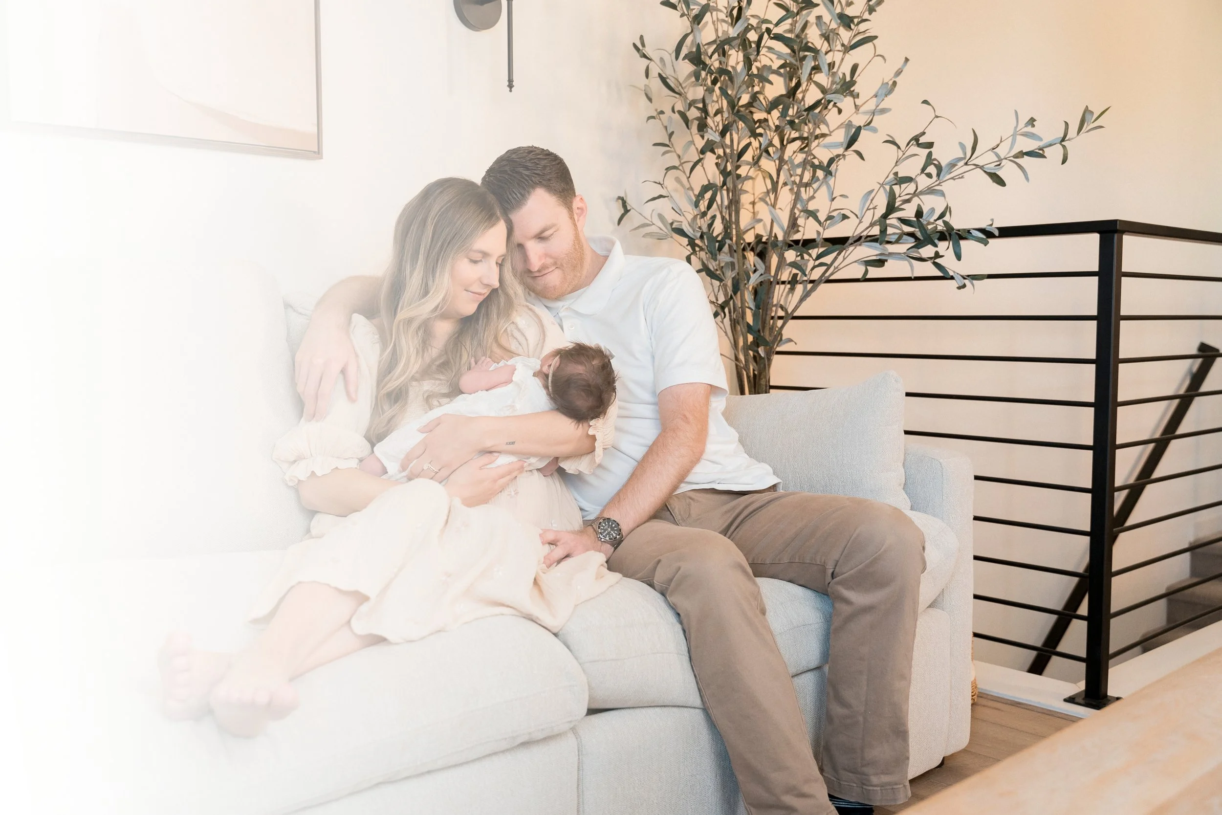 A family of three sitting on a light-colored couch, a woman holding a newborn baby, a man sitting beside her, all looking at the baby lovingly, in a cozy, modern living room with a large plant in the background.