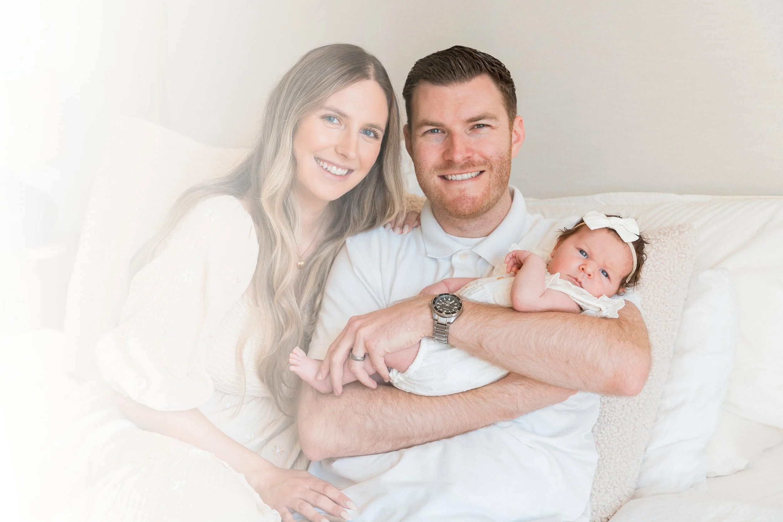A happy family with a woman, a man, and a baby girl, sitting on a white couch in a bright room.