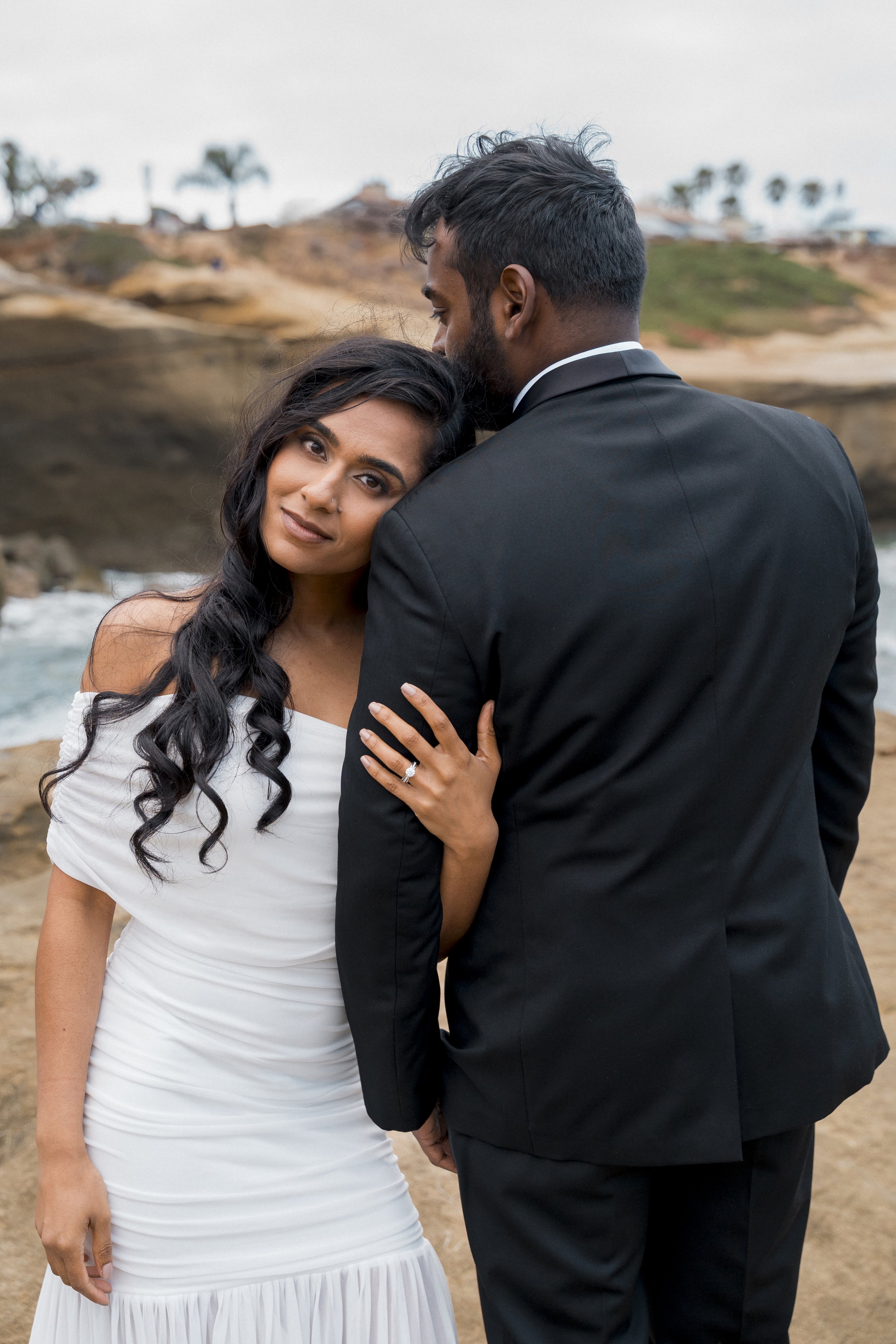 A couple in love standing face to face outdoors, smiling and looking into each other's eyes, with a historic church and palm trees in the background.