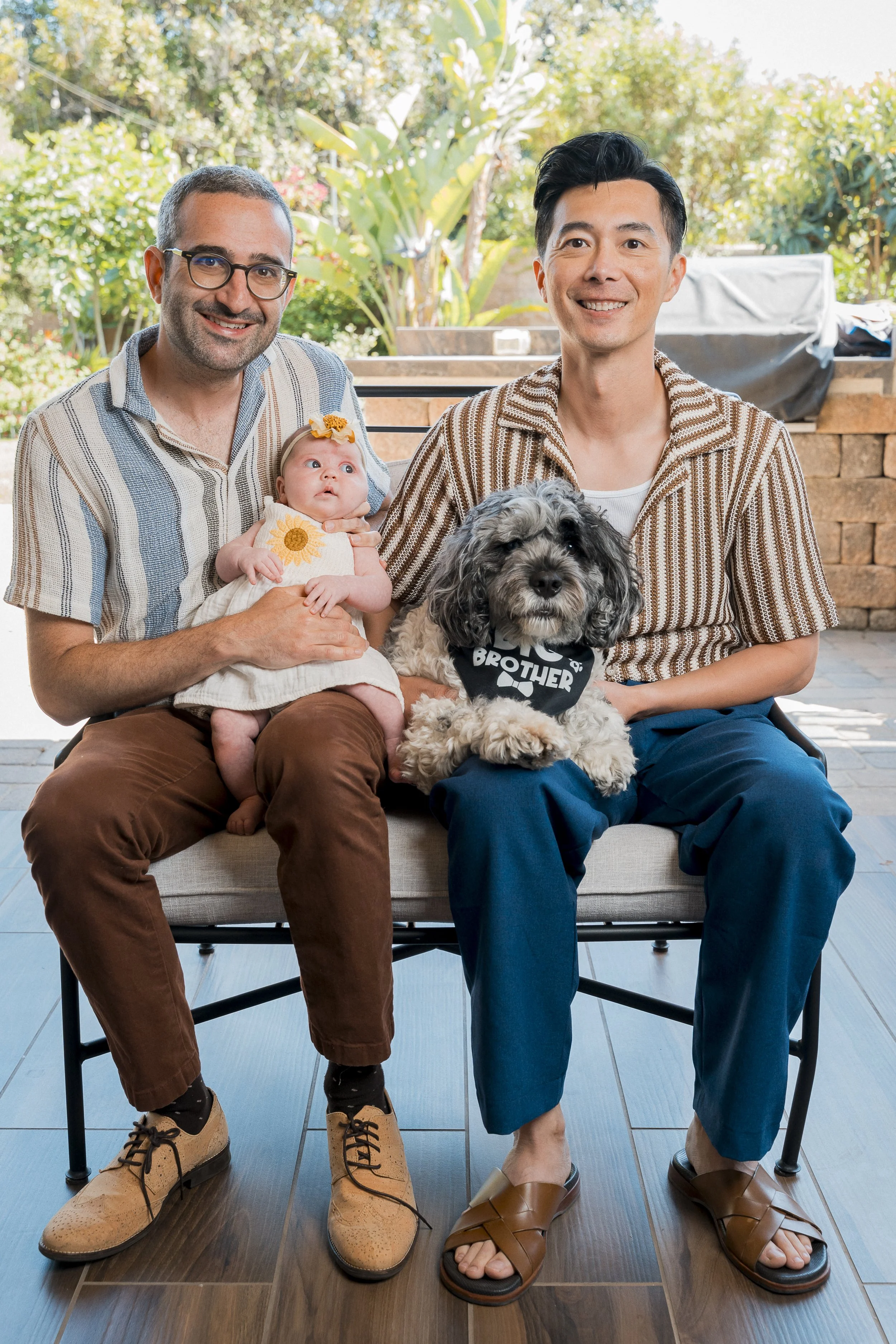Two men sitting on a bench, one holding a baby girl, with a dog on the men's lap. The background shows a garden with greenery.