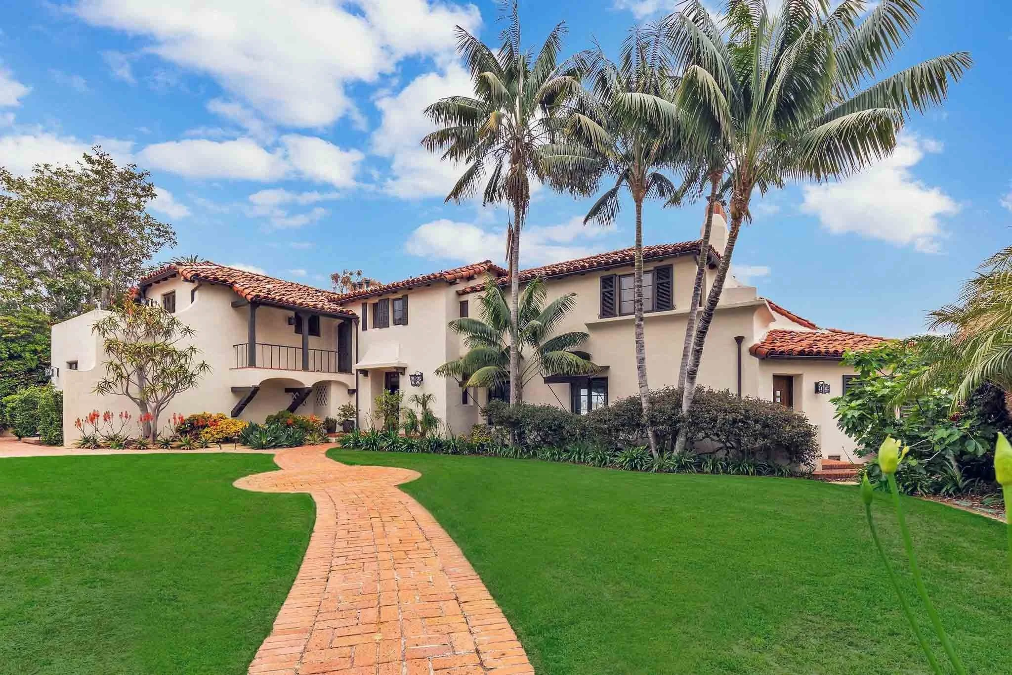 A house with a Mediterranean style, white walls, red tile roof, surrounded by palm trees, green lawn, and a brick pathway leading to the entrance under a blue sky with scattered clouds.