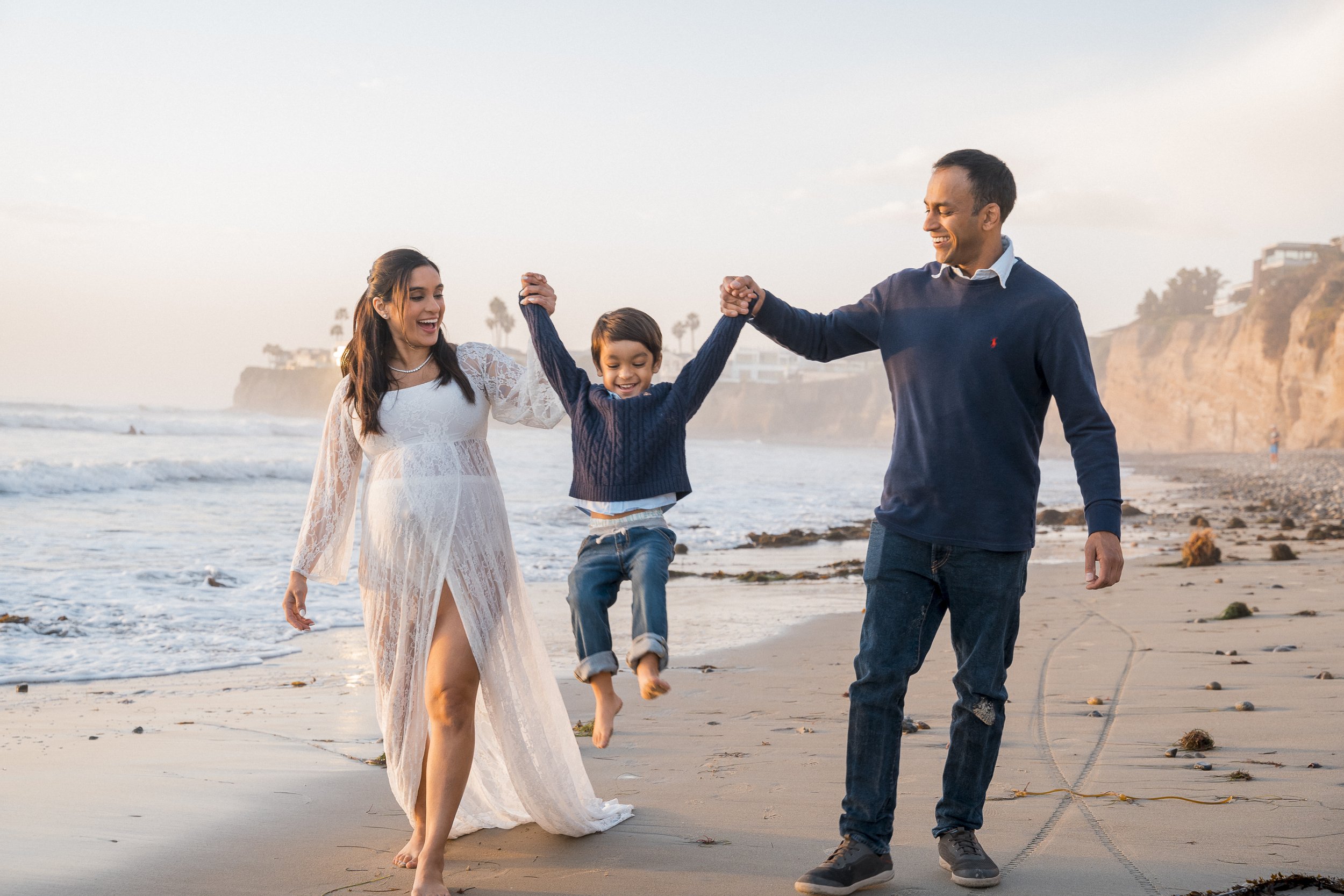 A family of three, a woman, a man, and a boy, playing and holding hands on a beach during sunset.