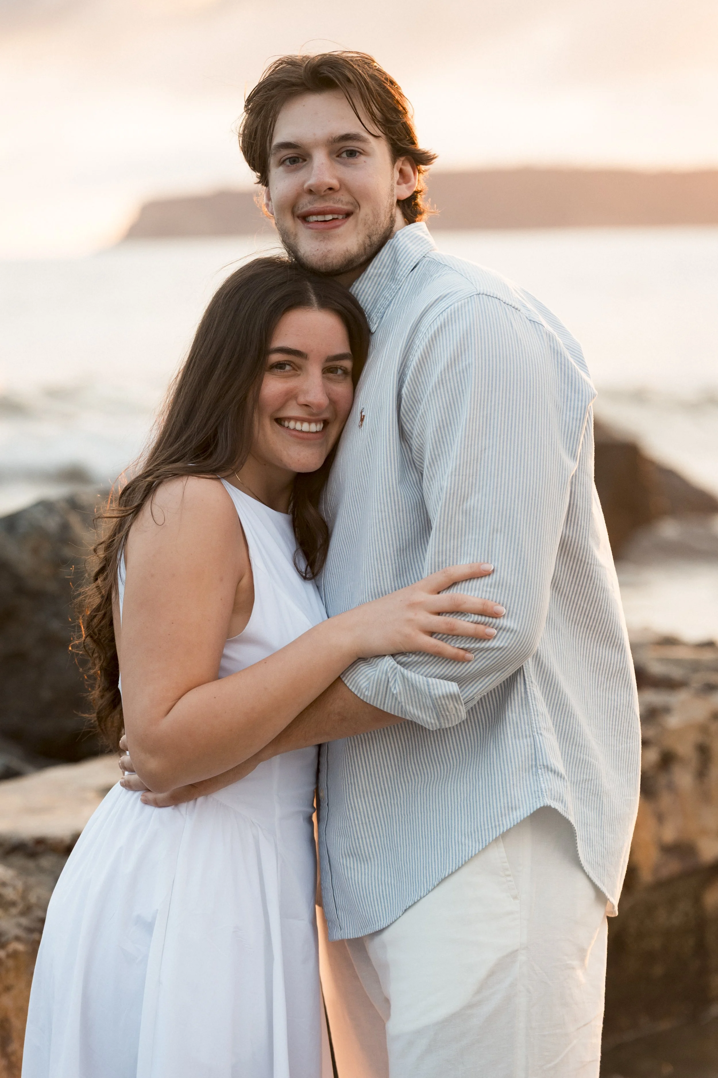 A couple dancing barefoot on the beach with waves in the background, enjoying a sunny day.