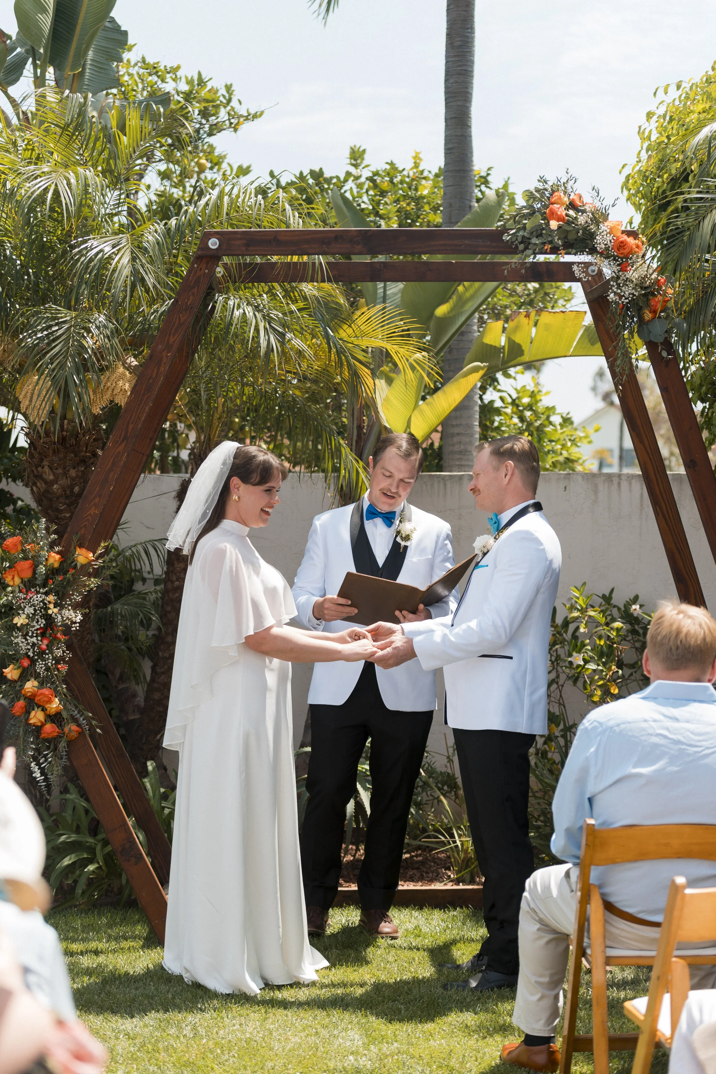 A bride in a white lace wedding dress and a groom in a blue suit, standing outside near green palm trees. The bride holds a bouquet of pink and white flowers with feathers, and they share an intimate, happy moment.