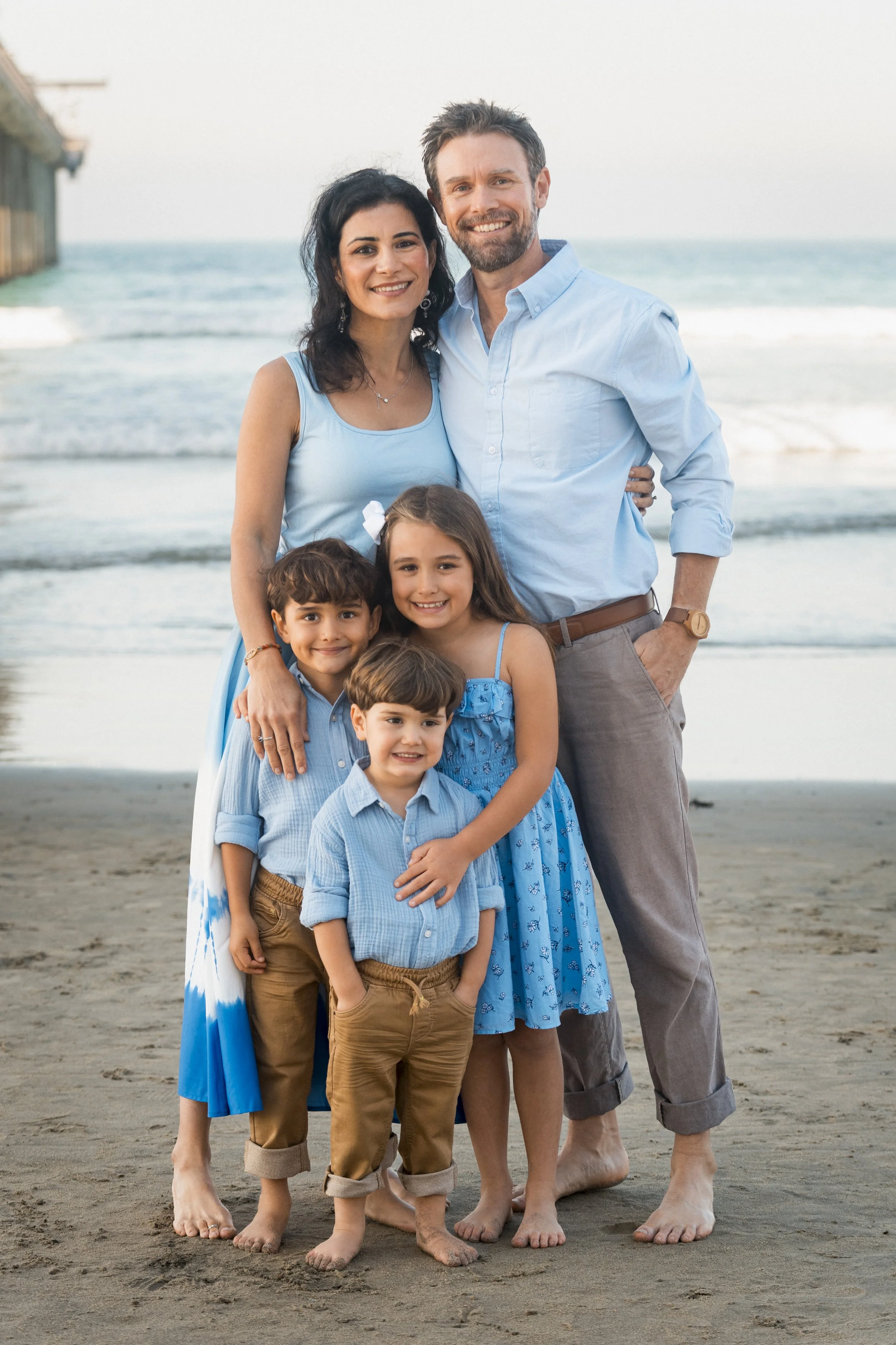 Family of five on the beach, posing together, smiling, in casual blue clothing with the ocean and a pier in the background.