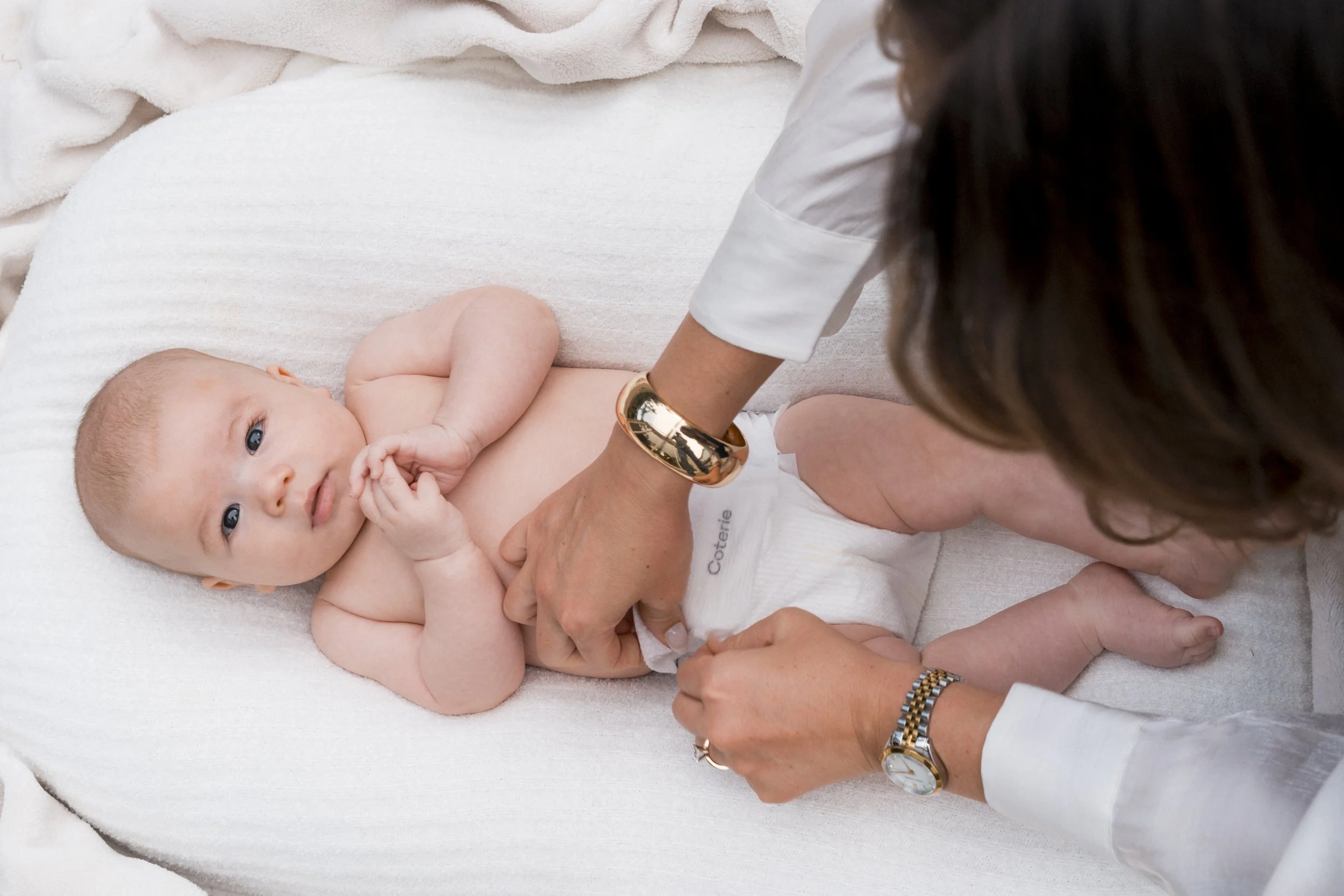 A baby lying on its back on a white changing pad, looking up. An adult woman, possibly the mother, is adjusting the baby's diaper. The woman is wearing a white shirt, a gold bracelet, a watch, and a ring. The baby is only wearing a diaper, and the background includes soft white fabric.