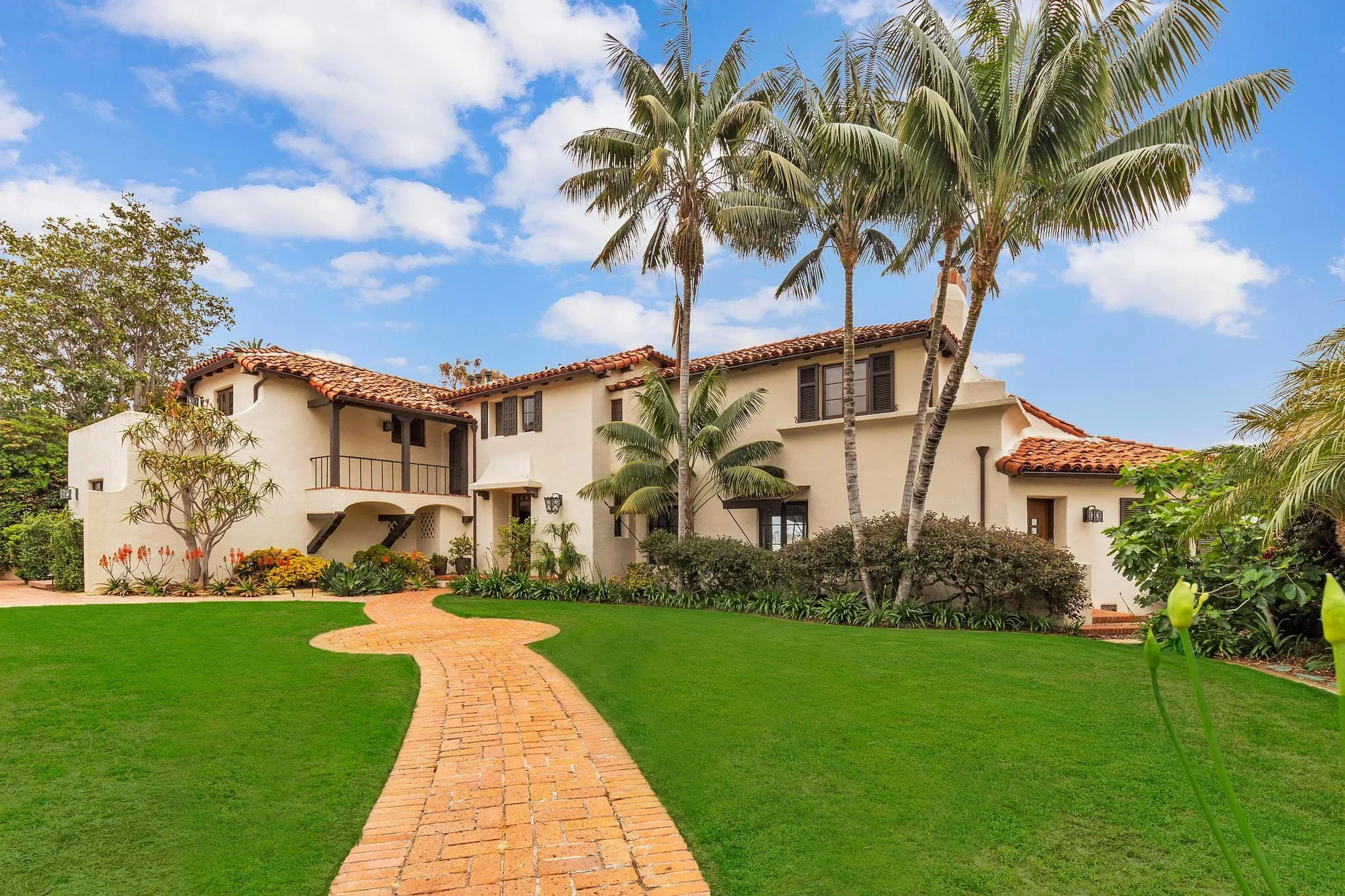 A house with a Mediterranean style, white walls, red tile roof, surrounded by palm trees, green lawn, and a brick pathway leading to the entrance under a blue sky with scattered clouds.
